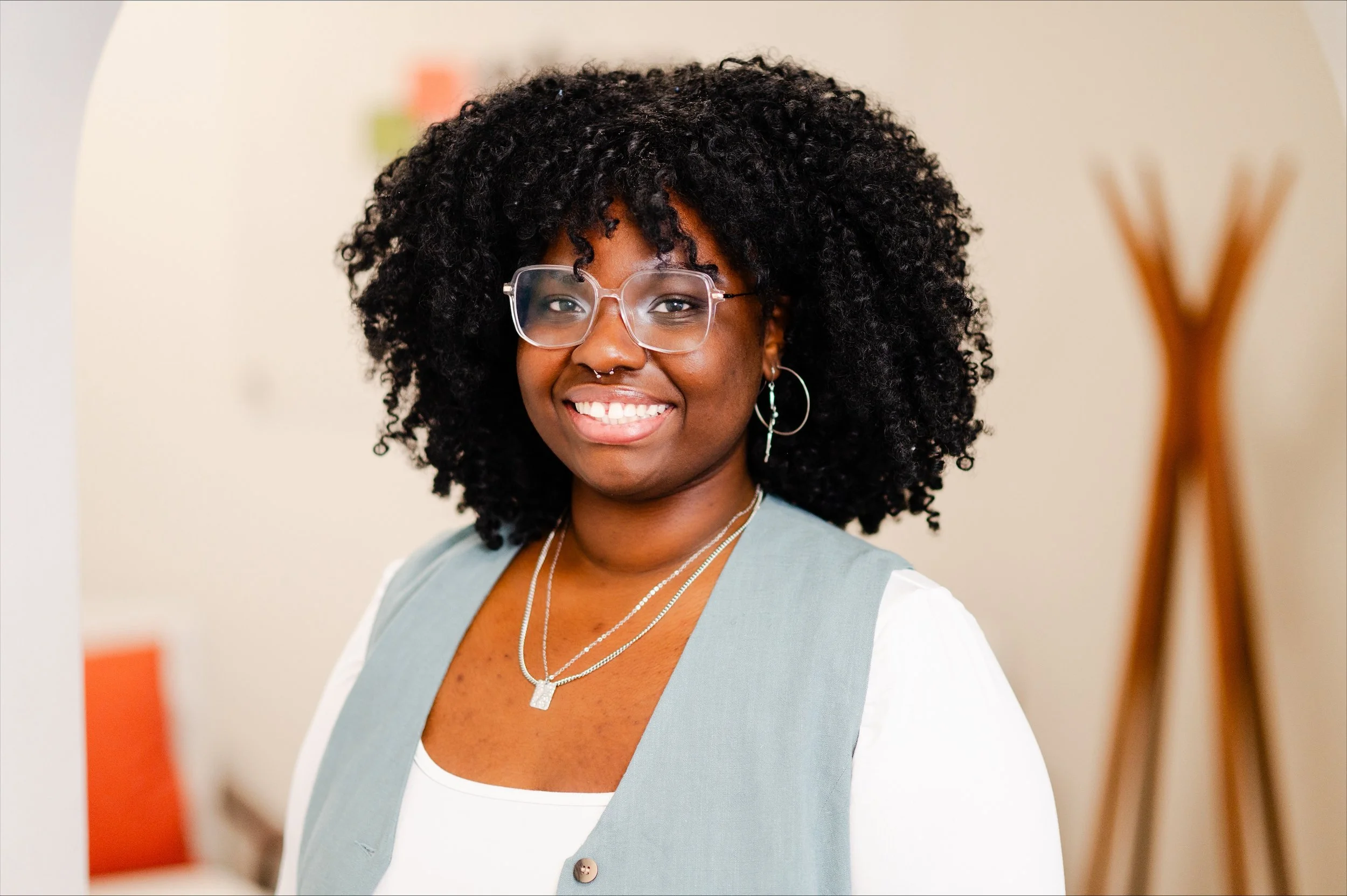 A smiling woman with curly black hair, wearing glasses, layered necklaces, a septum piercing, earrings, a light green vest over a white top, and standing in a bright room with a blurred background.