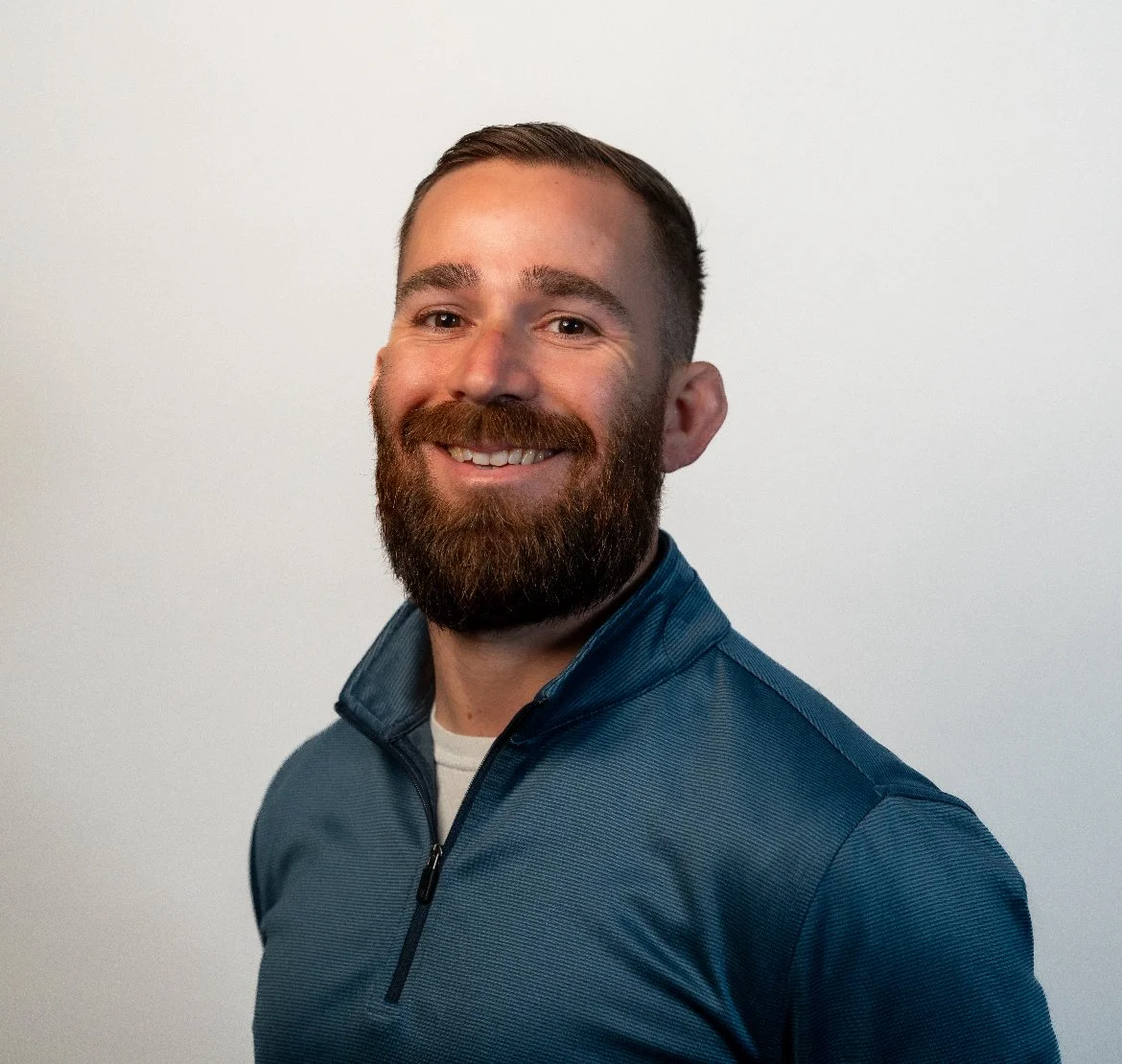 Portrait of a smiling man with a beard wearing a blue zip-up jacket against a light background.