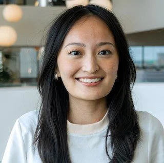A young woman with long black hair smiling, wearing a white top, indoors with large windows in the background.