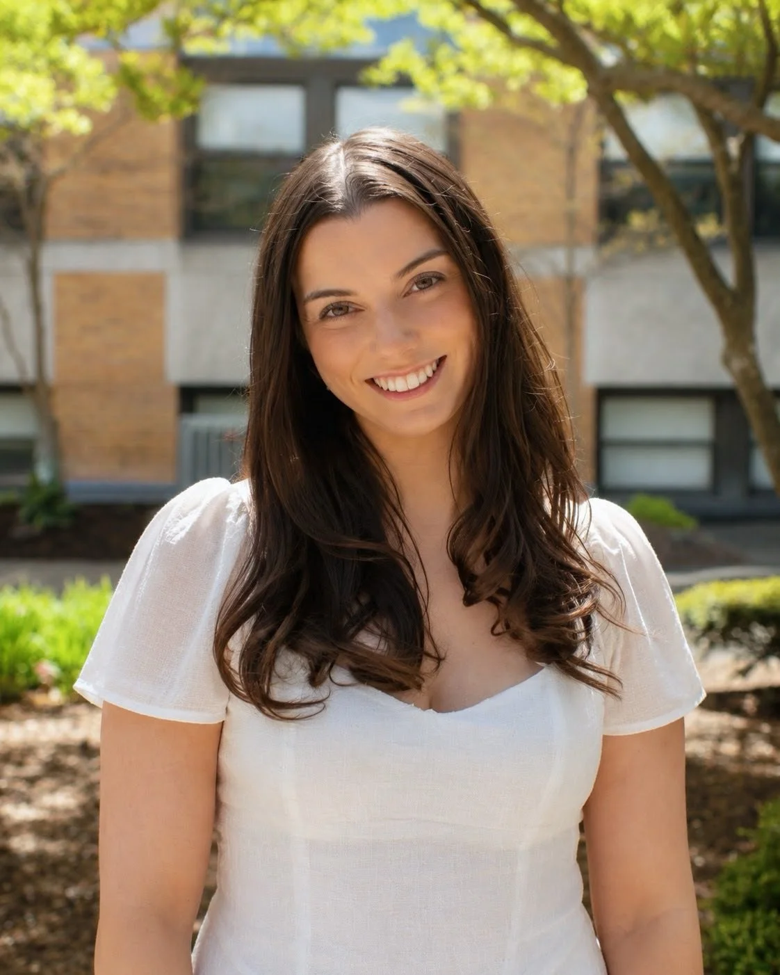 A young woman with dark hair and a white top smiling outdoors with trees and a building in the background.