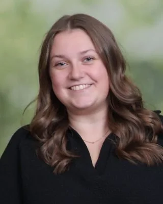 A young woman with long, wavy brown hair, smiling, wearing a black top, with a blurred green outdoor background.