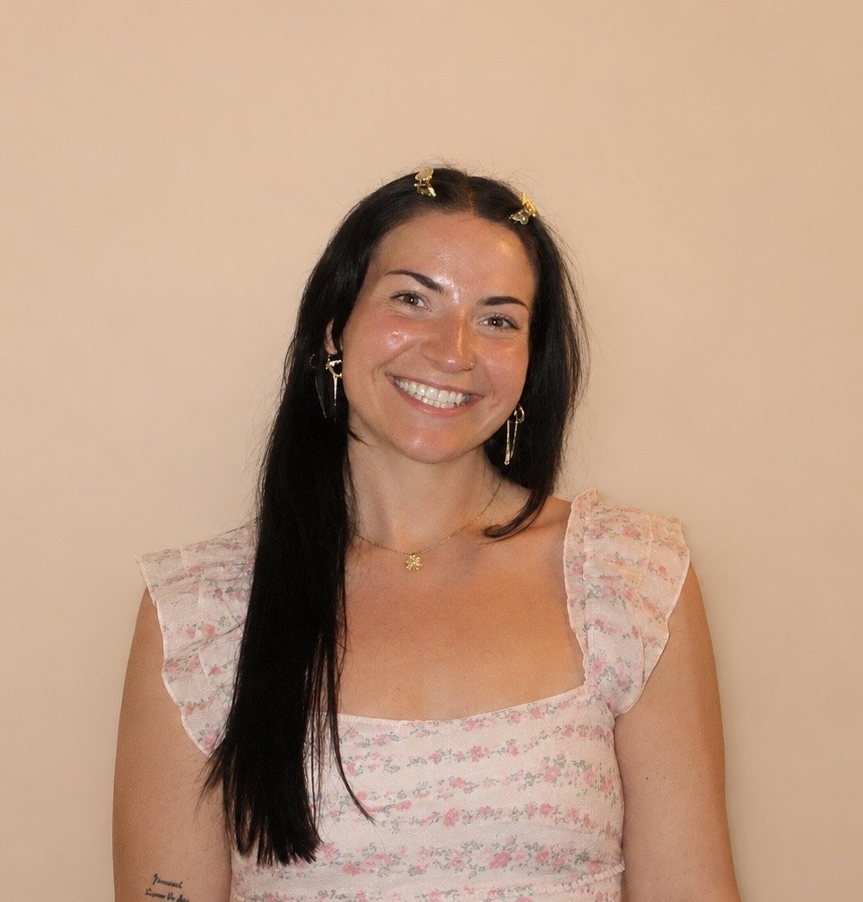 A woman with long black hair smiling, wearing a pink floral dress, gold earrings, a gold necklace, and hair clips, standing against a plain beige wall.
