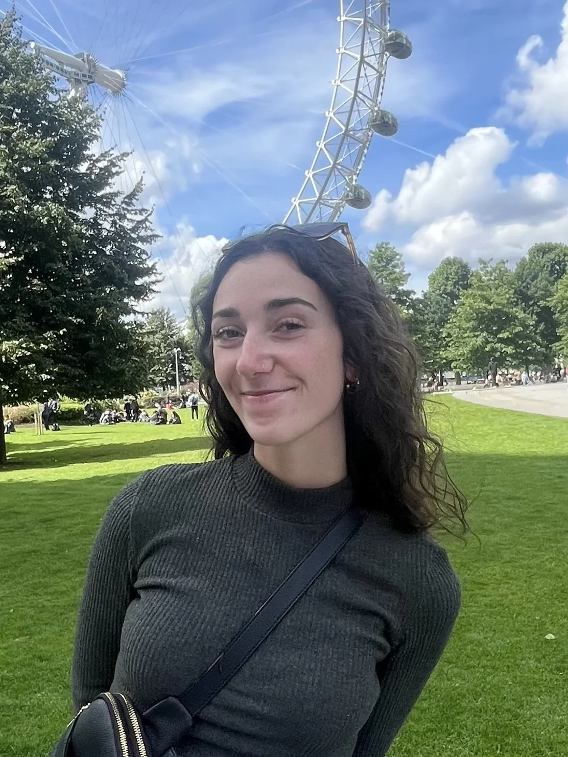 A woman with dark wavy hair and wearing a dark ribbed sweater, smiling outdoors in a park with green grass, trees, and a large Ferris wheel in the background under a partly cloudy sky.