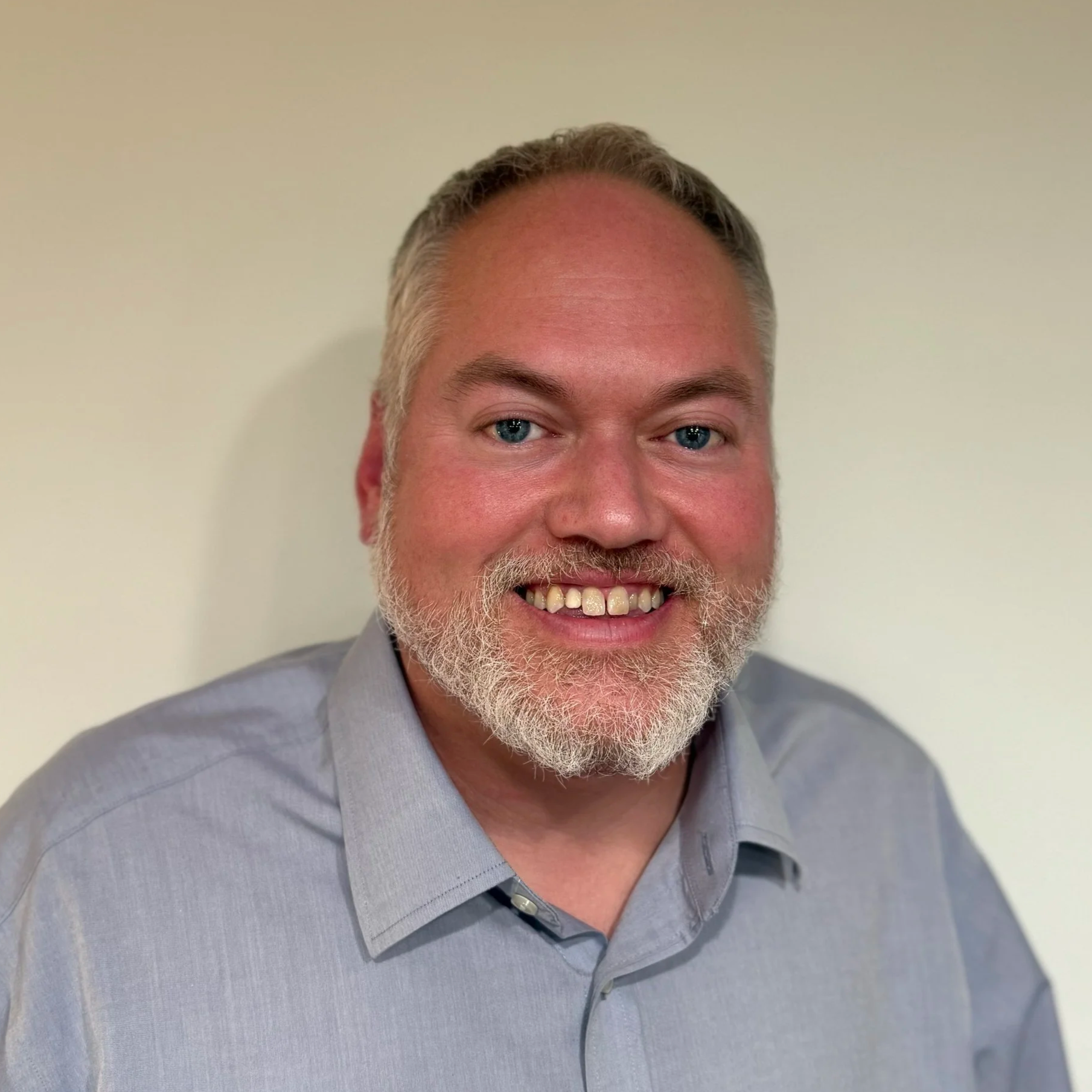Close-up of smiling man with gray hair, beard, blue eyes, wearing a light gray dress shirt, standing against a beige wall.