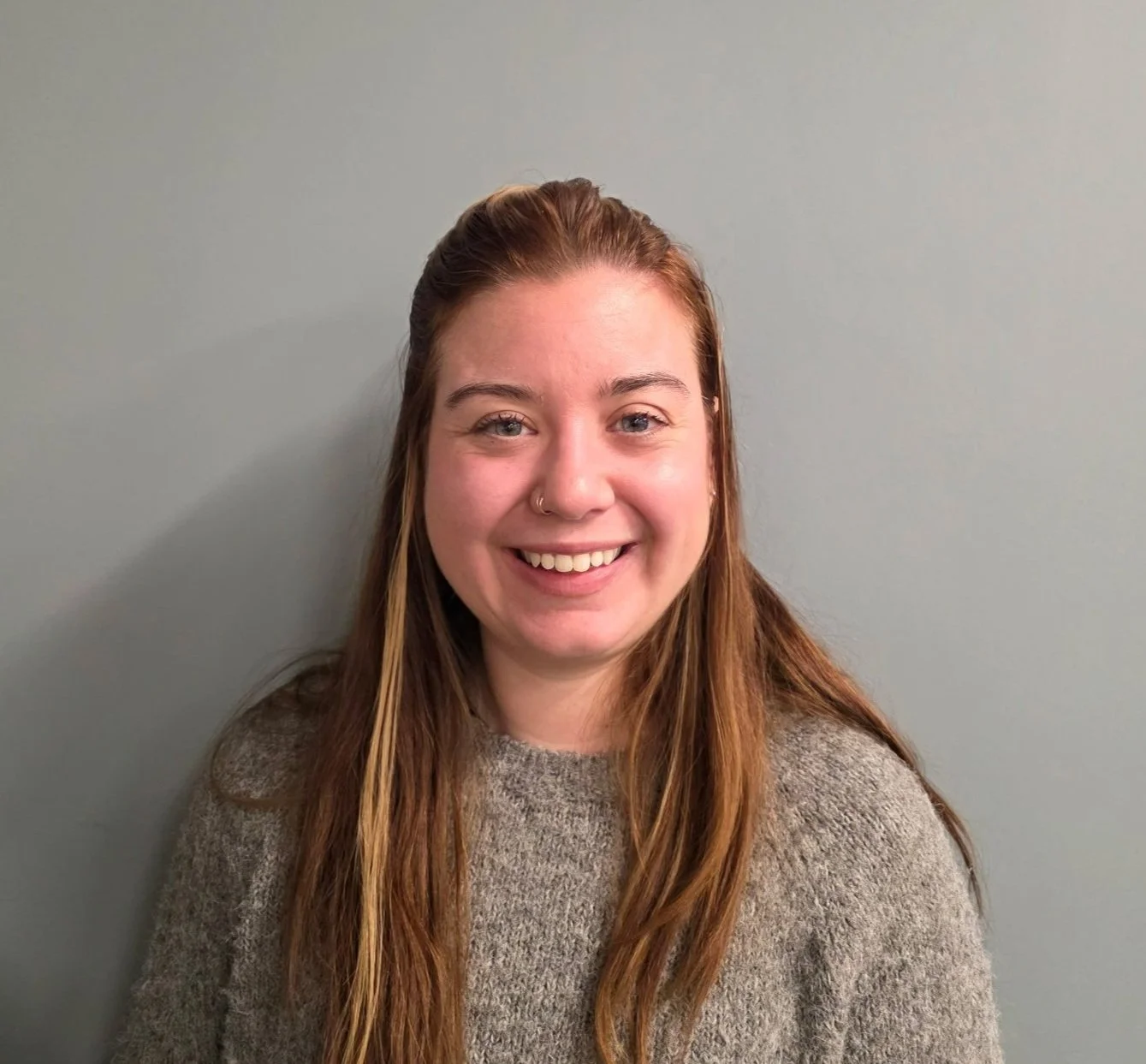 A young woman with long, reddish-brown hair smiling at the camera, wearing a gray sweater, standing against a plain gray wall.