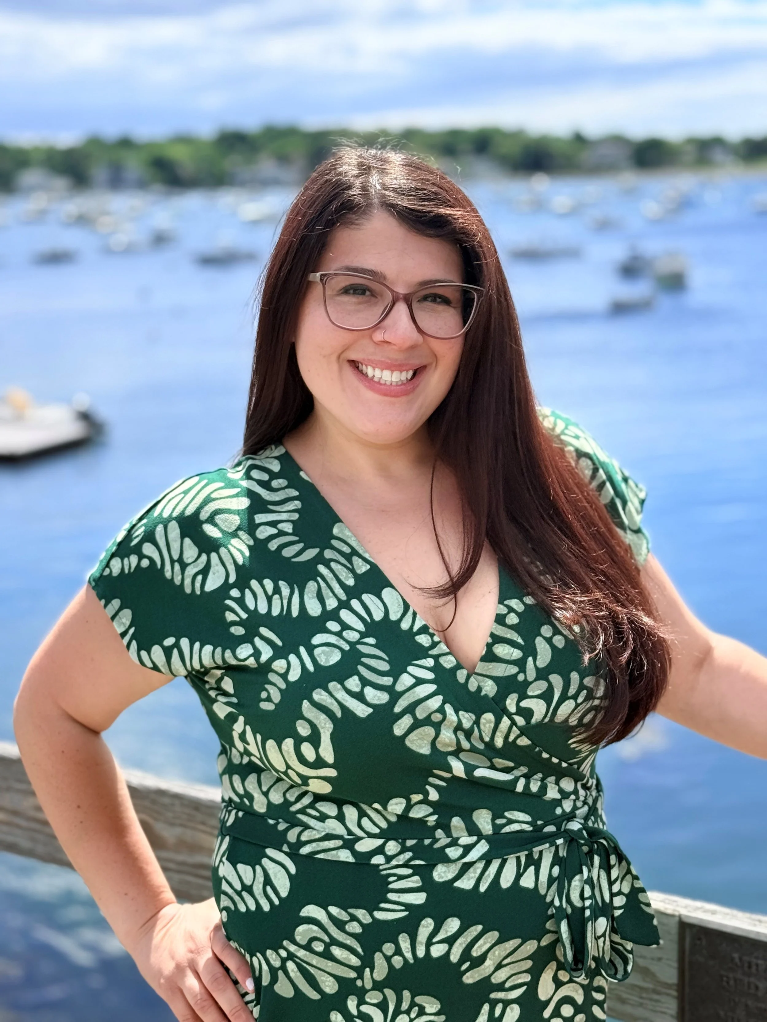 A woman with long brown hair wearing glasses and a green dress with a white leaf pattern, smiling outdoors near a body of water with boats and a distant shoreline with trees.