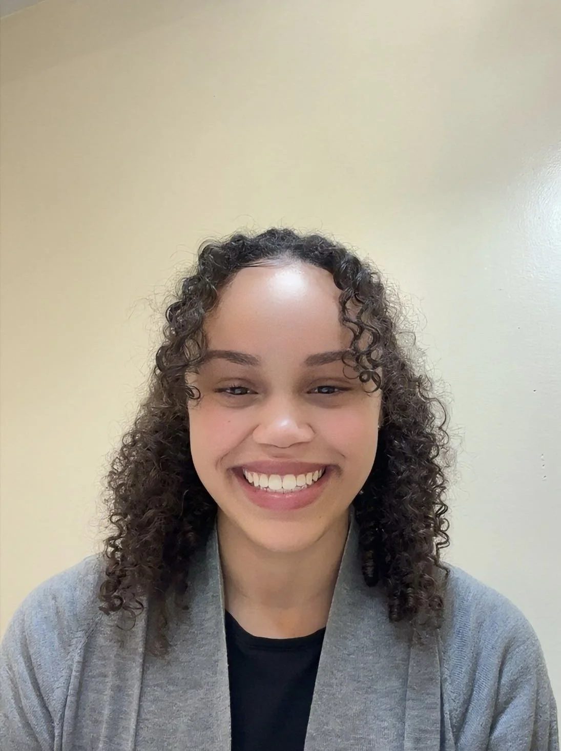 Smiling woman with curly hair, wearing a gray jacket and black shirt, standing against a plain light-colored wall.