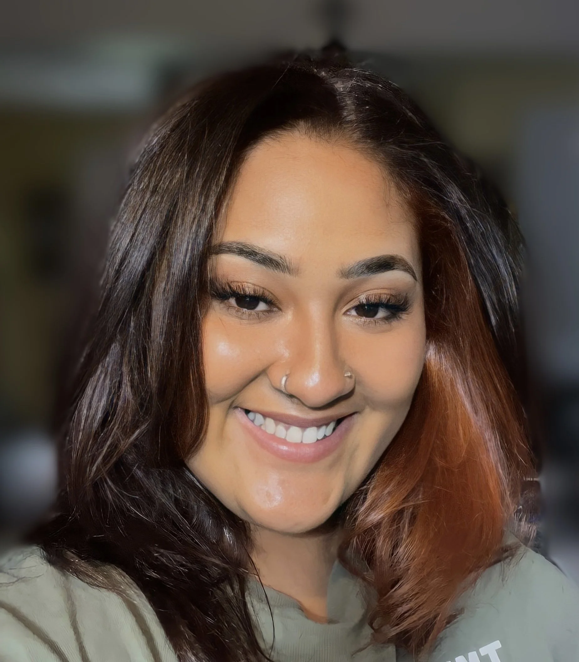 A woman with long dark brown hair, smiling showing her teeth, wearing a light-colored top.