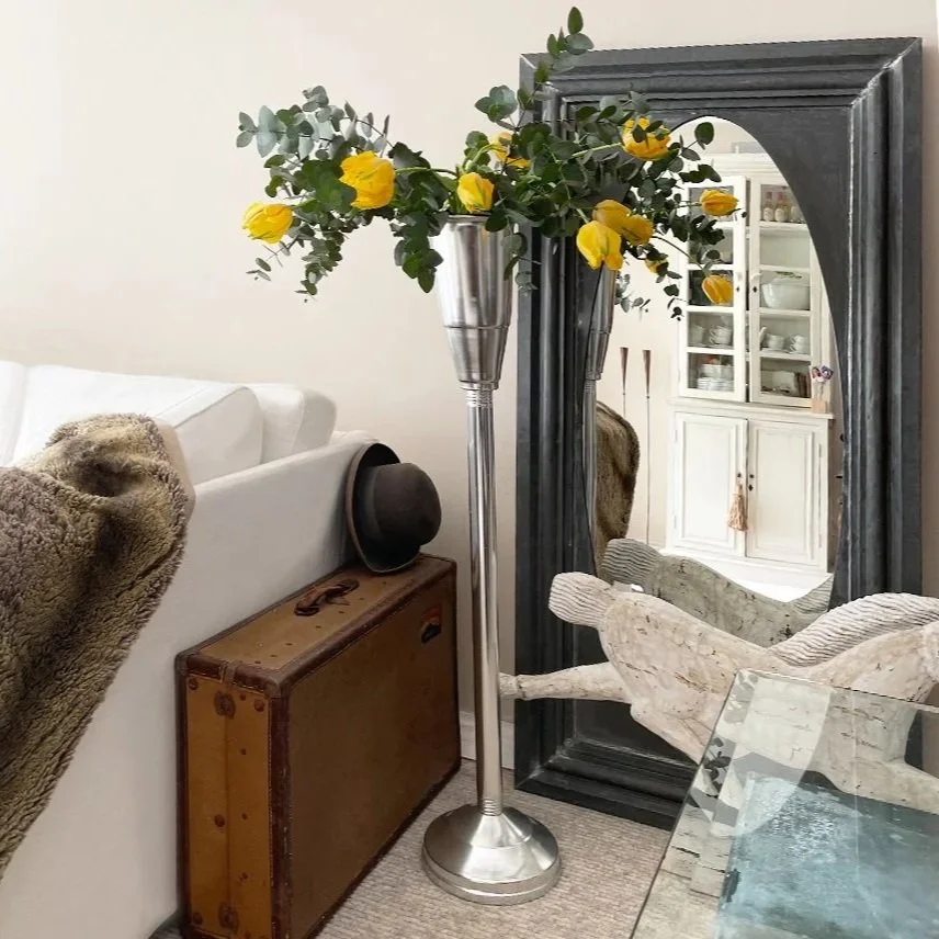A narrow living room with a white sofa, a vintage suitcase, a black hat, a silver vase with yellow flowers and greenery, a large black framed mirror, a wooden sculpture, and a glass table.