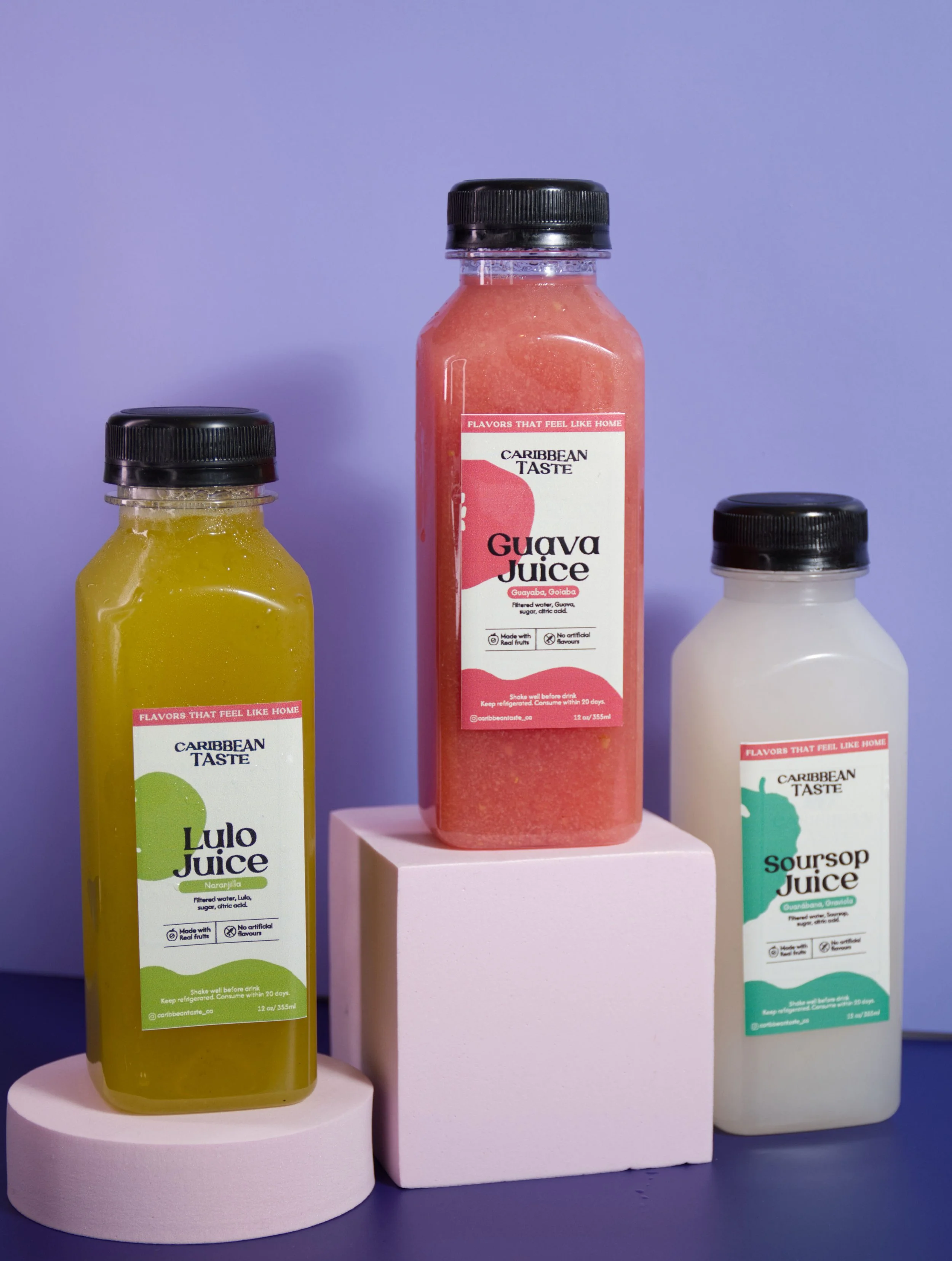Three bottles of Caribbean fruit juices against a purple background, with the guava juice in the center on a pink block, and the lulo juice and soursop juice on the sides on white blocks.