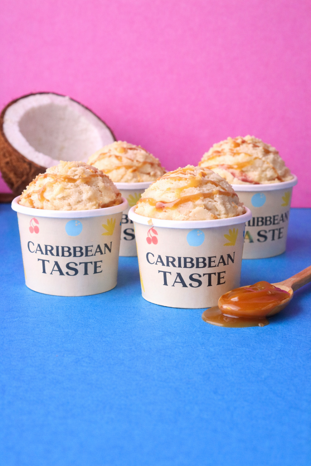 Four cups of ice cream with caramel drizzle and chopped nuts, with a halved coconut in the background and caramel sauce on a spoon in the foreground, on a blue surface with a pink background.