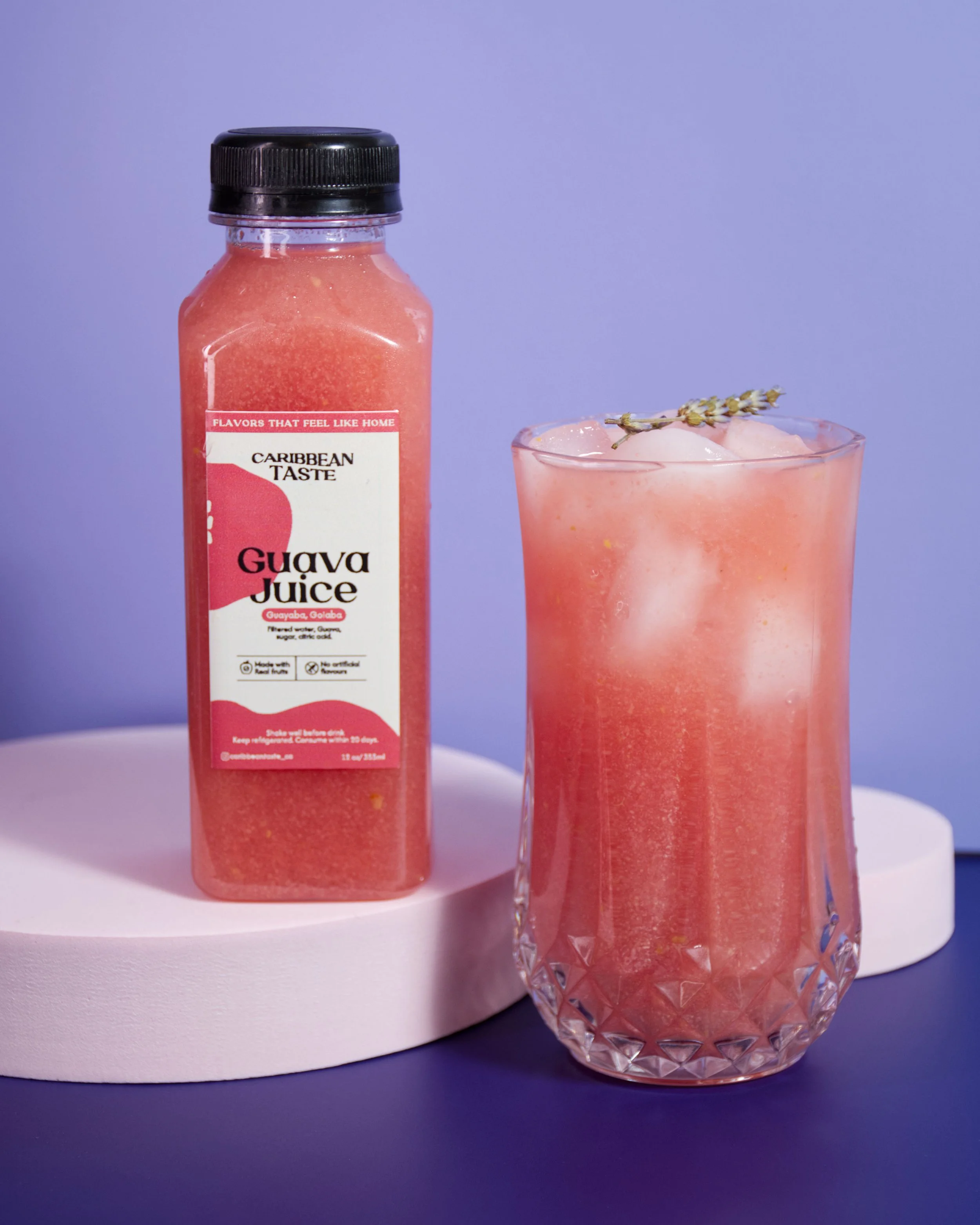 Bottle of Caribbean Taste Guava Juice next to a glass of guava juice with ice and a sprig of lavender on top, on a pastel pink and purple background.