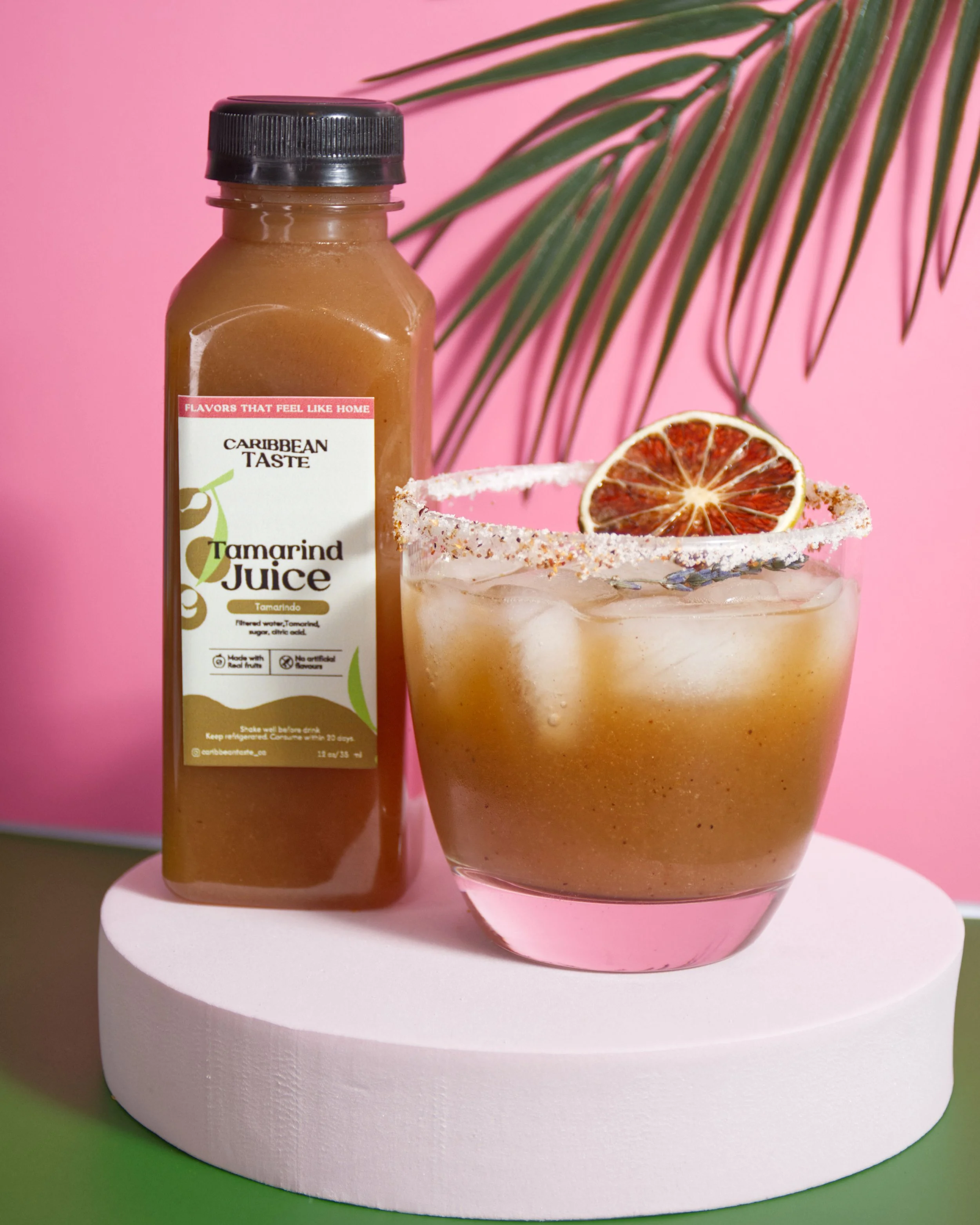 A bottle of Caribbean Tamarind juice and a glass of tamarind drink garnished with a dried lime slice and a salted rim, placed on a white circular platform against a pink background with green palm leaves.