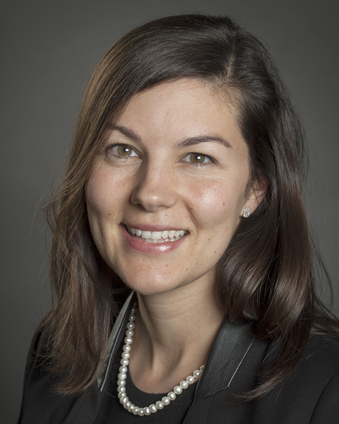 Headshot of a woman with shoulder-length brown hair, wearing a black blazer, pearl necklace, and pearl earrings, smiling against a gray background.