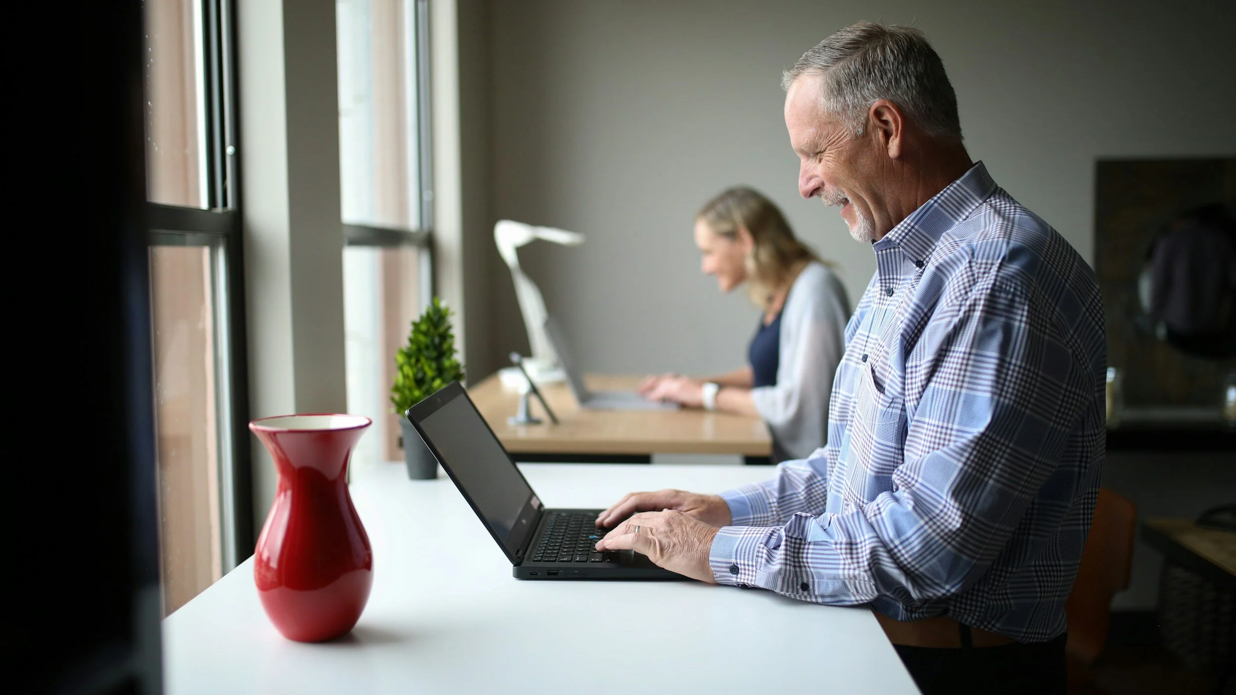 Elderly man working on a laptop at a white desk, with a woman working on her laptop in the background, and a red vase and small plant on the desk.