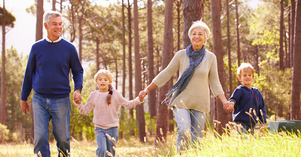 Older couple holding hands with two young children, walking in a forest clearing on a sunny day.