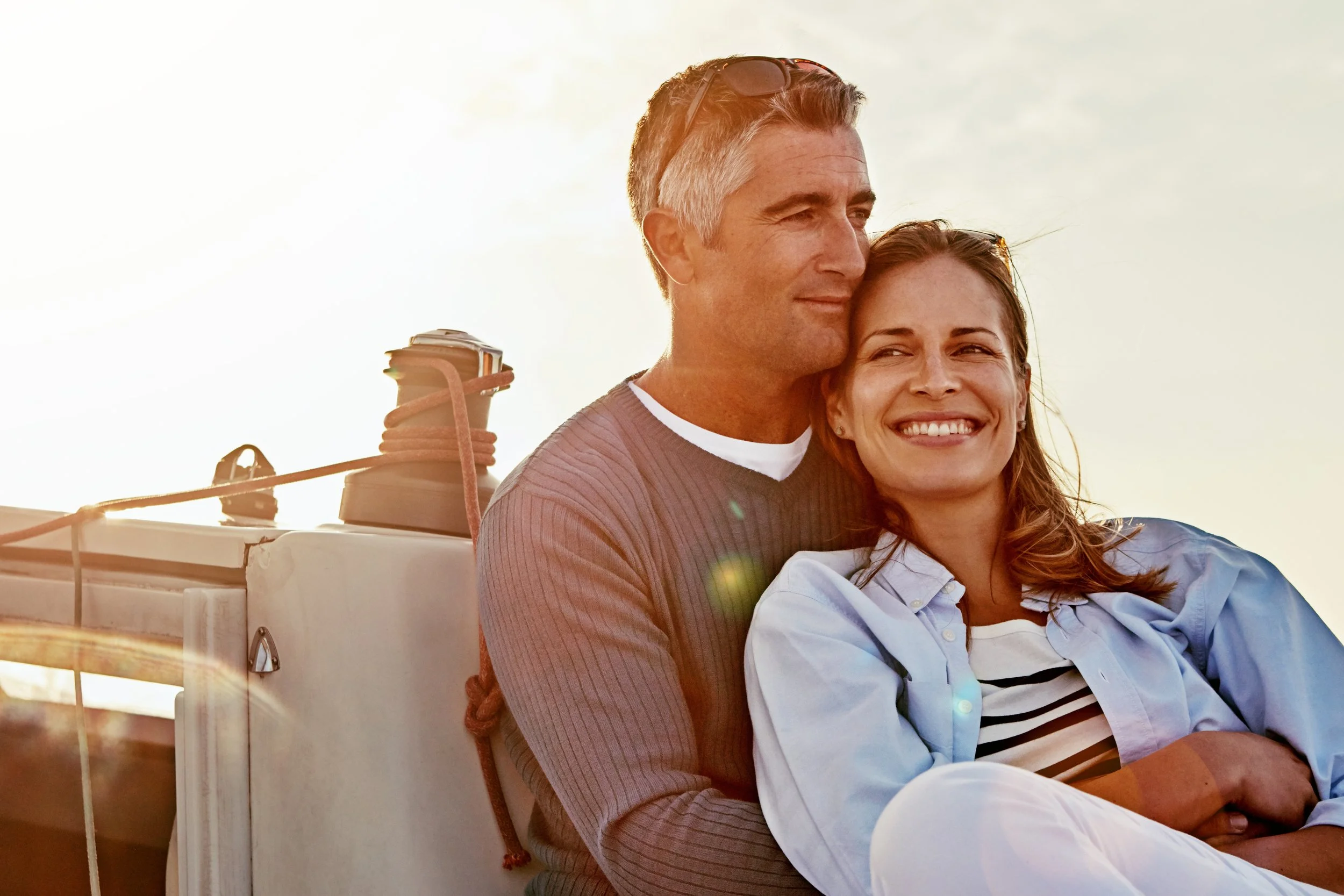 A man and woman sitting close together on a boat, smiling, with the sun or bright sky behind them.