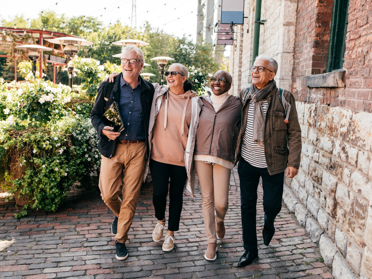 Four diverse friends walking together outside, smiling and enjoying each other's company on a cobblestone street near a brick building with greenery and outdoor heaters in the background.
