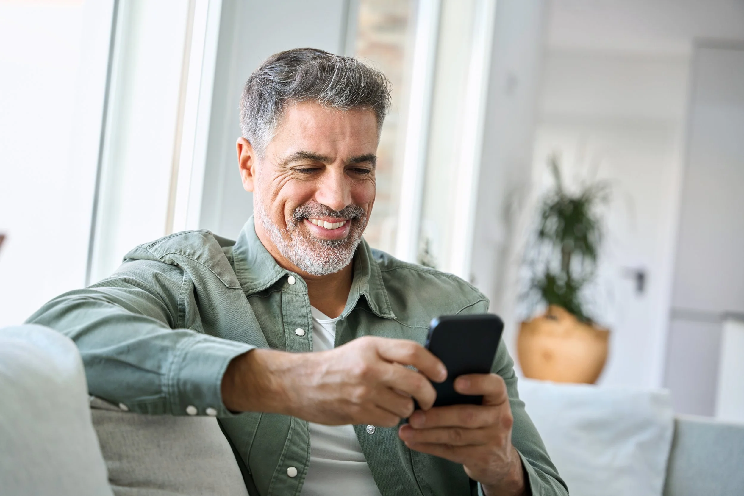 A middle-aged man with gray hair and a beard smiling while looking at his smartphone in a bright living room.