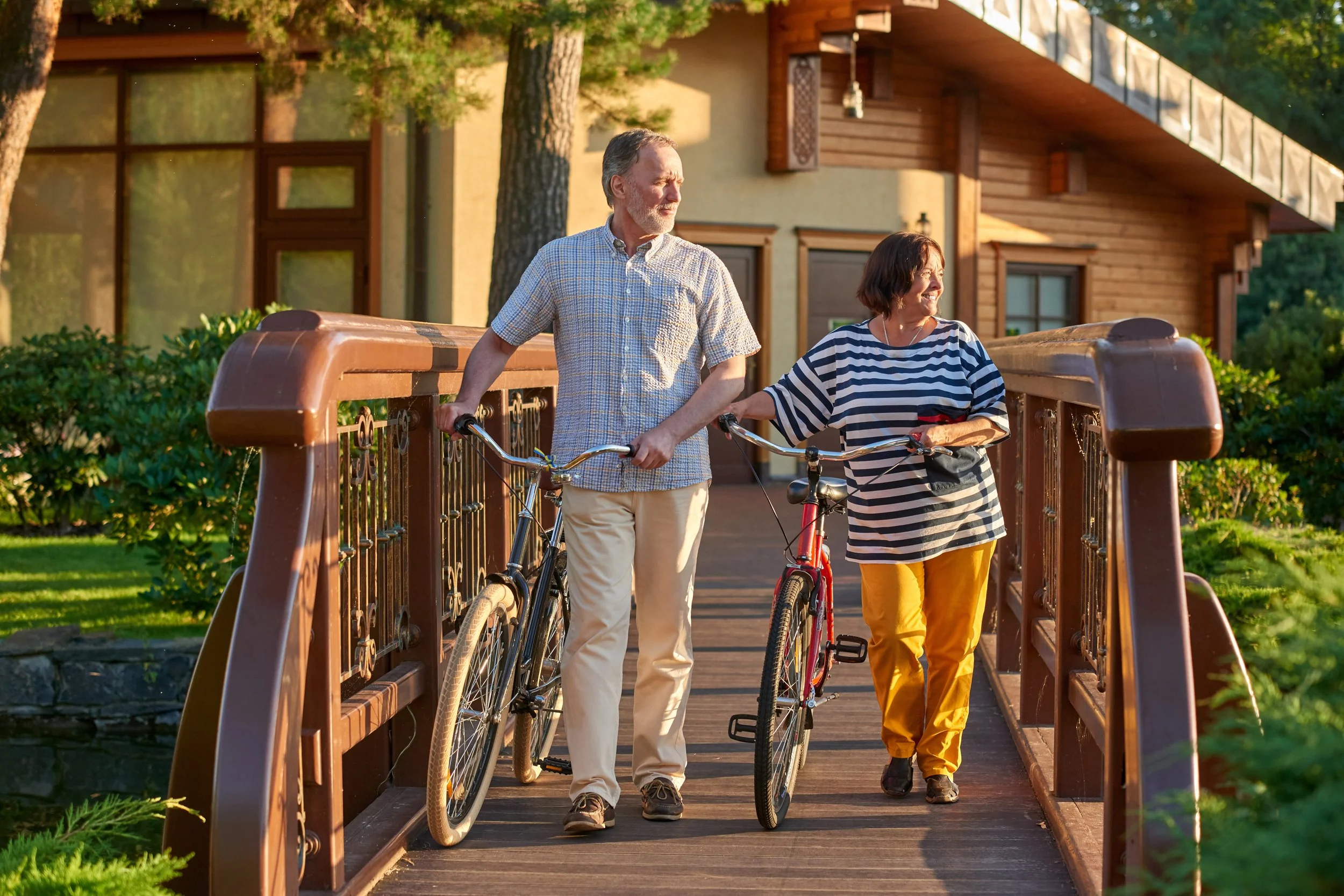 A senior couple walking on a wooden bridge holding bicycles, smiling, with greenery and a house in the background during sunset.