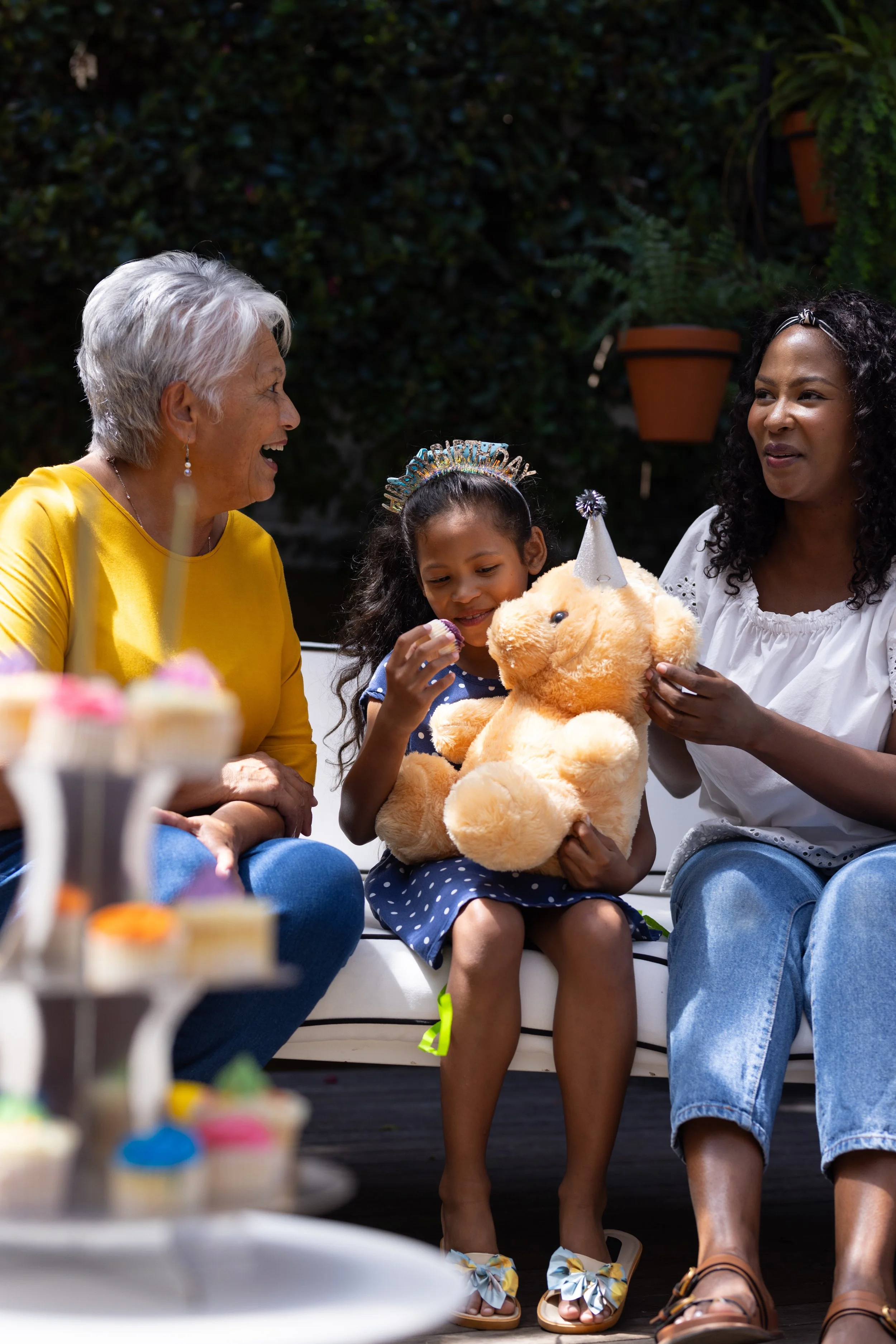A woman wearing a yellow shirt, a young girl with a birthday headband holding a teddy bear, and another woman on the right sitting together outdoors at a birthday party, with cupcakes visible in the foreground.
