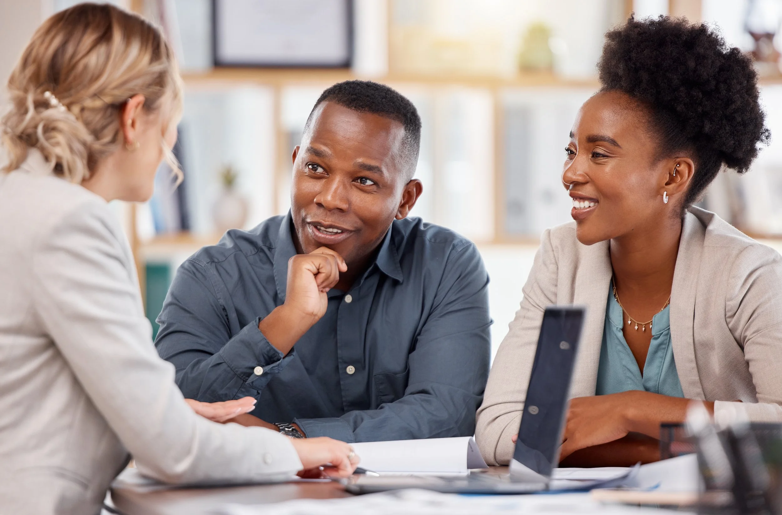 Three diverse colleagues in a business meeting, engaged in conversation around a table, with a laptop and documents present.