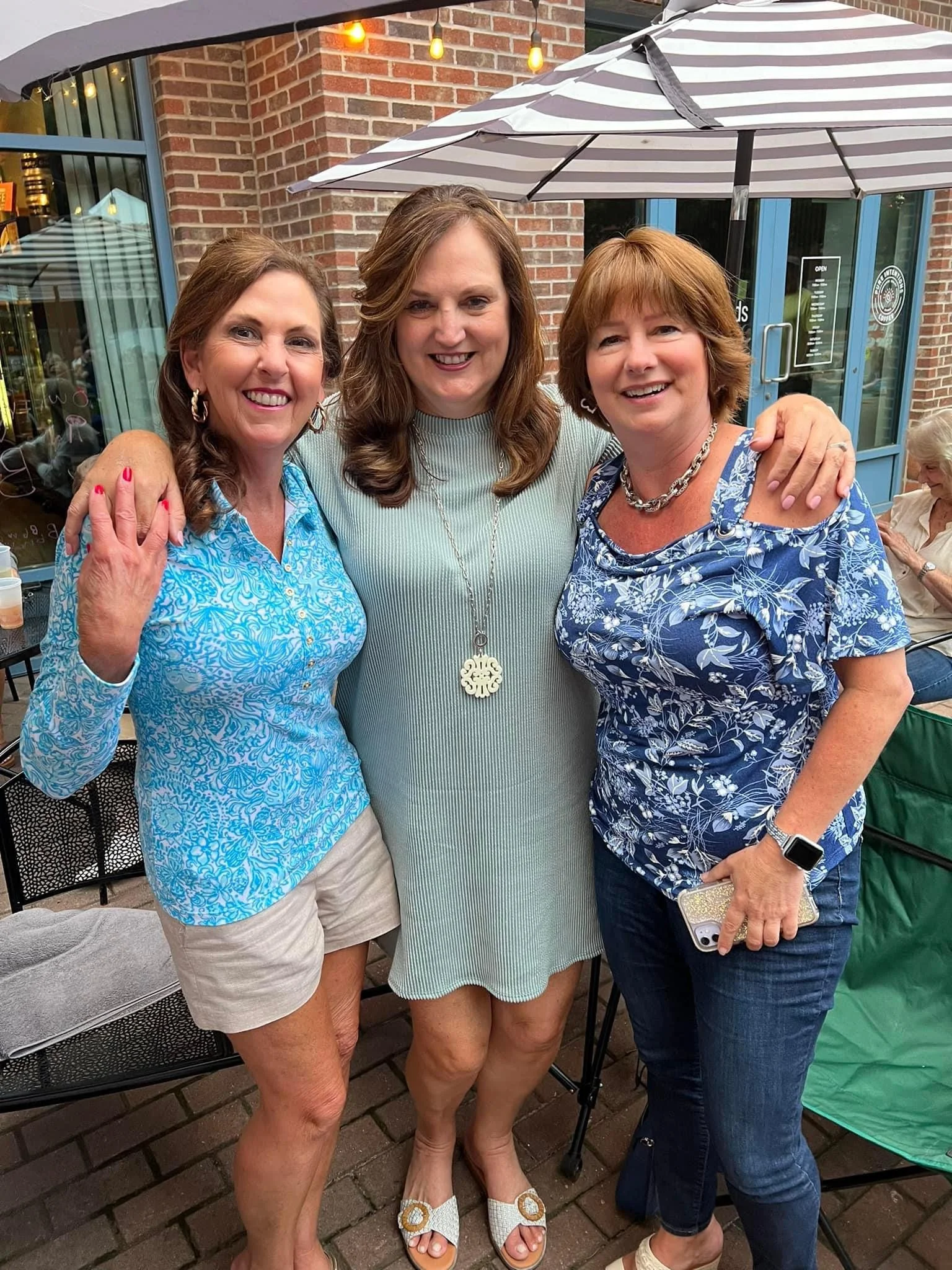 Three women standing outside under an umbrella, smiling and posing for a photo, with a brick building in the background.