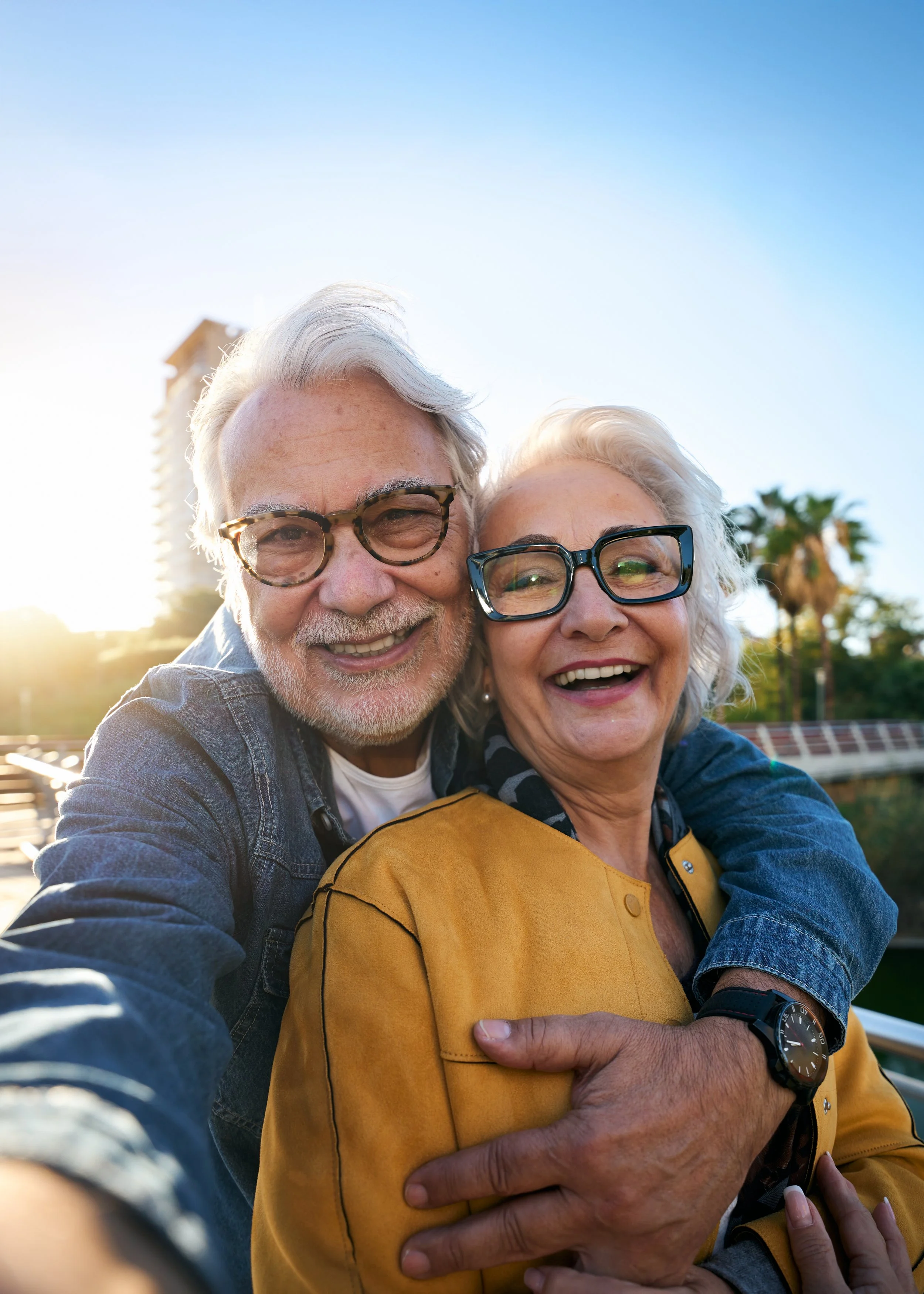 Two elderly people, a man and a woman, smiling happily and taking a selfie outdoors during sunset, with palm trees in the background.