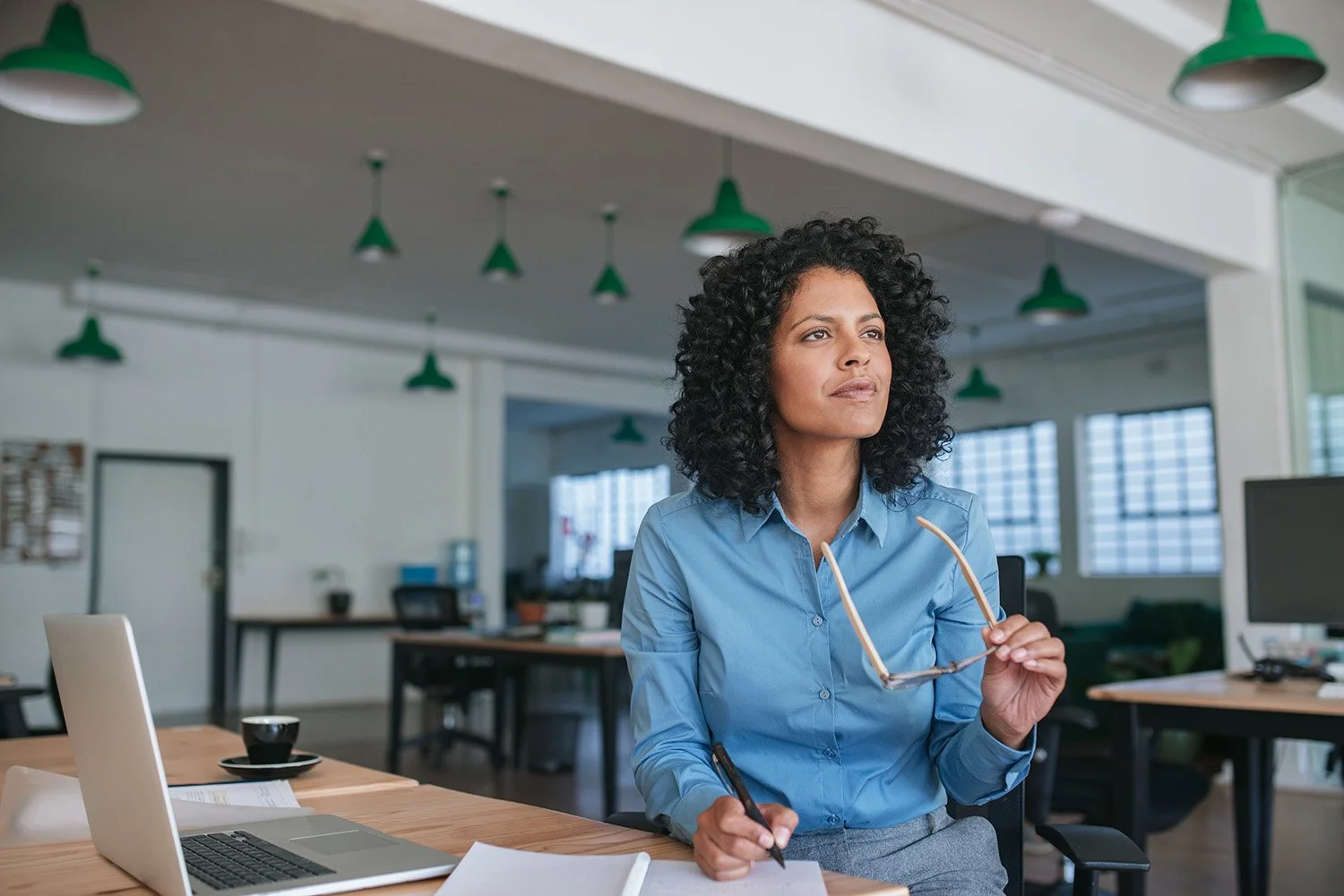 A woman with curly black hair wearing a blue shirt sits at a desk in an office, holding glasses in one hand and a pen in the other, looking thoughtfully into the distance.