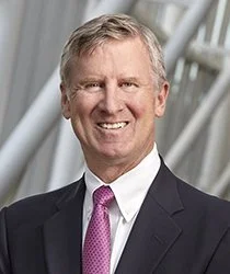 A professional man in a dark suit, white shirt, and pink tie, smiling against a modern architectural background.
