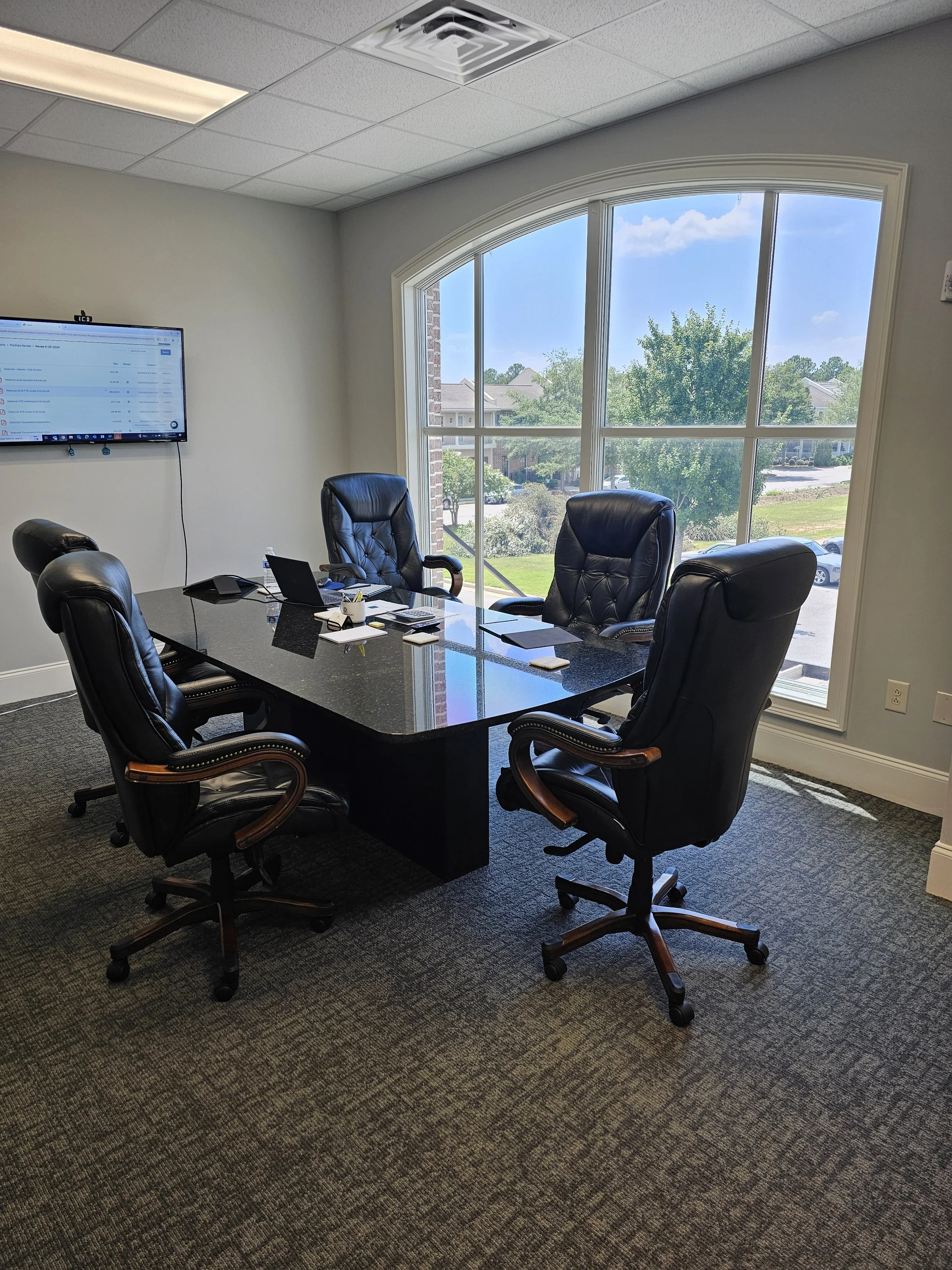 Empty conference room with five black leather chairs around a black marble table, large window showing trees and buildings outside, wall-mounted monitor on the left, carpeted flooring, and part of ceiling with recessed lighting.