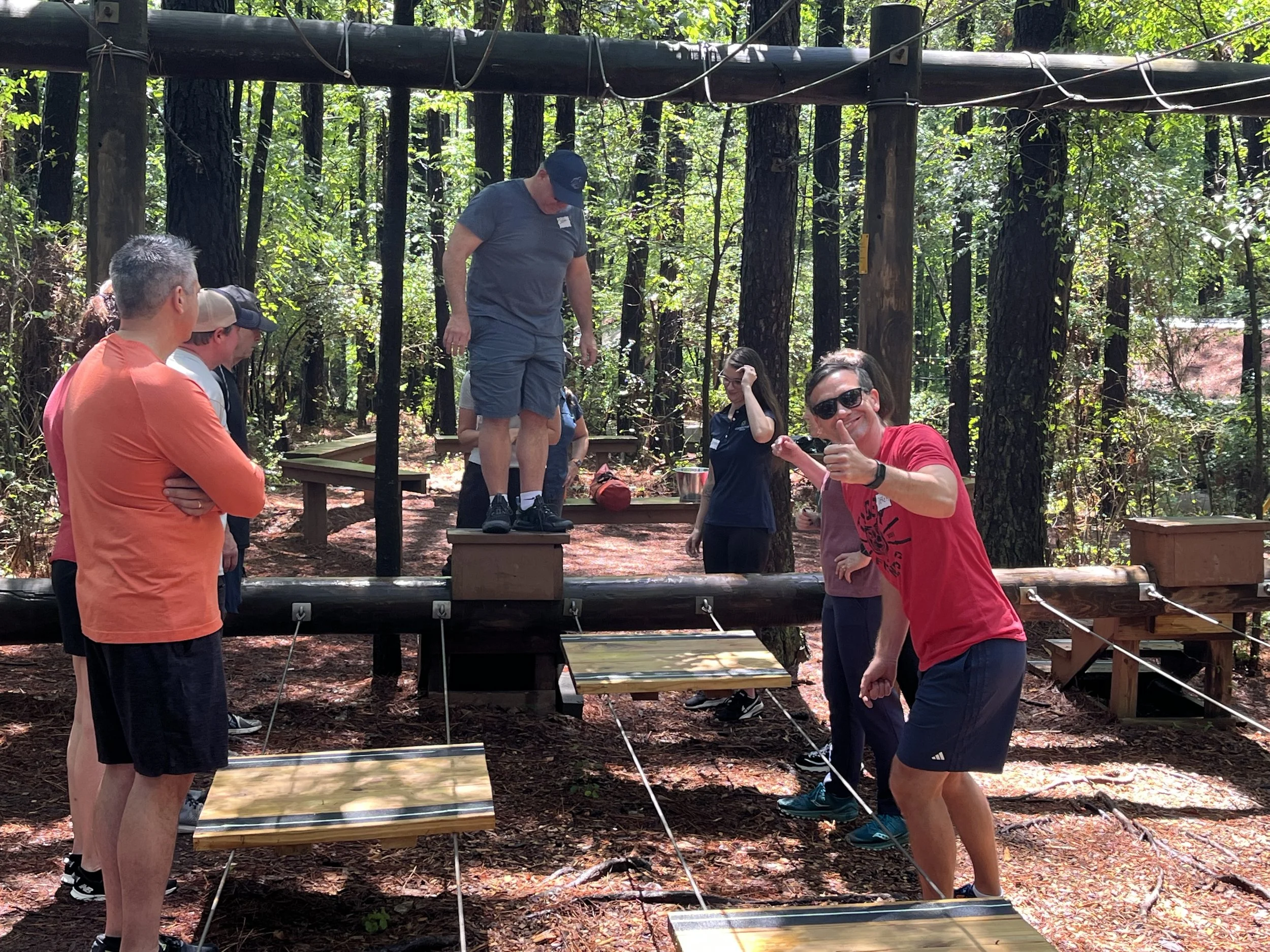Group of people in a forested outdoor obstacle course, with one person balancing on a raised wooden platform while others watch and smile, some giving thumbs up.