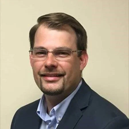A man with glasses, short brown hair, and a goatee smiling, wearing a navy blazer and light blue dress shirt, standing against a beige wall.