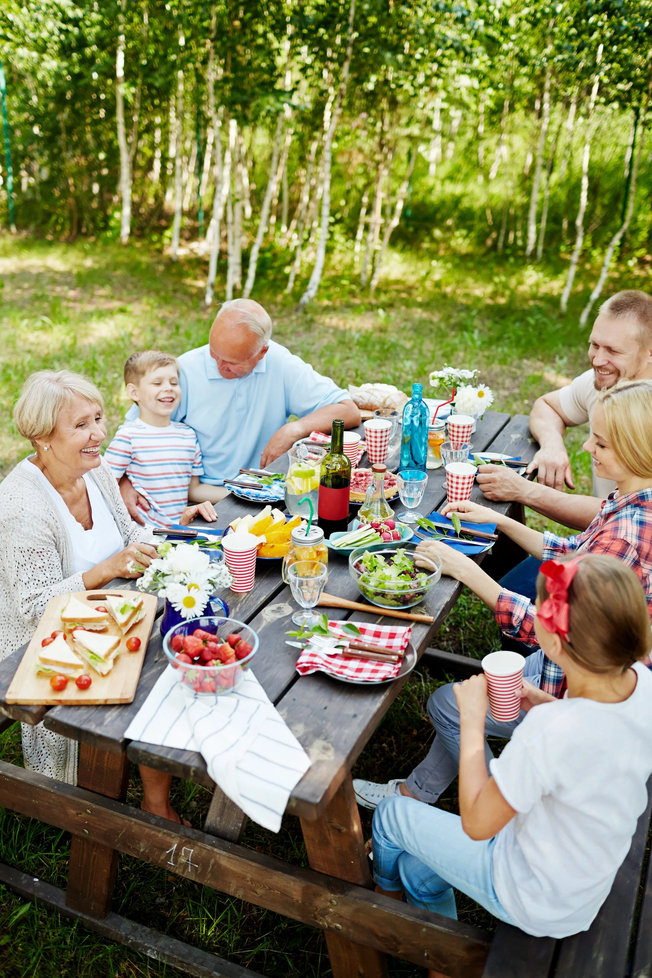A multi-generational family having a picnic at a wooden table outdoors surrounded by trees, with various foods, drinks, and flowers on the table.