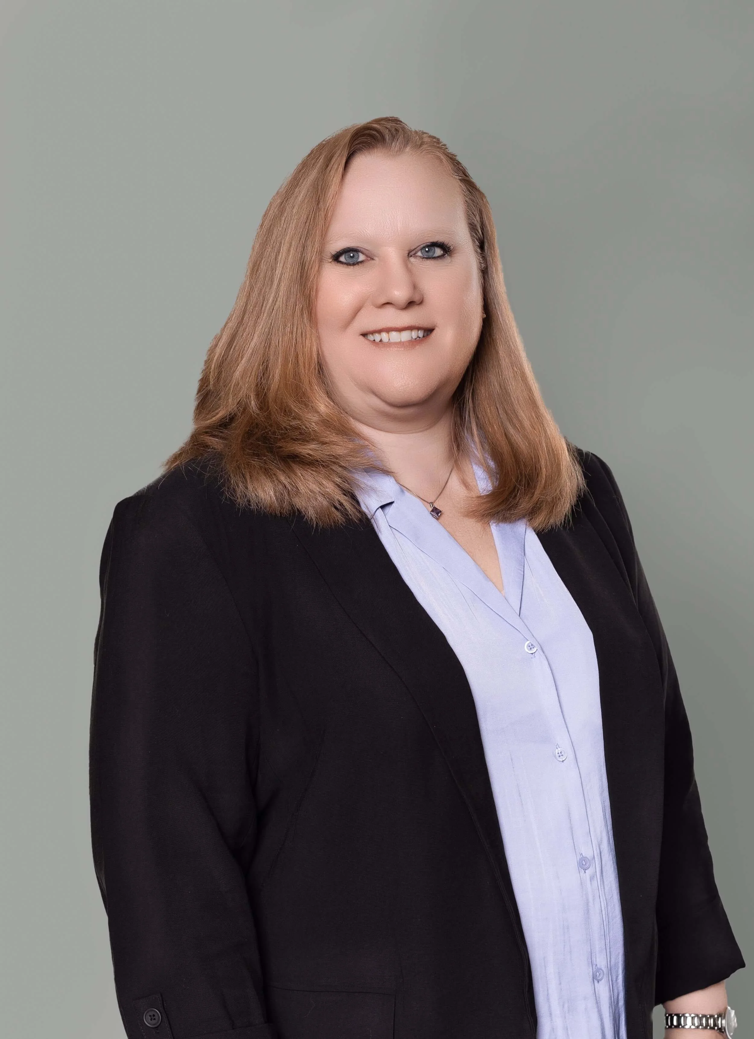 A professional woman with shoulder-length red hair, wearing a black blazer over a light blue button-up shirt, smiling against a plain light grey background.