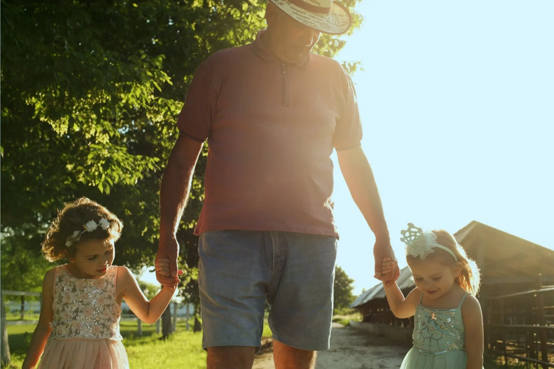 A man holding hands with two young girls, one in a pink dress and another in a light green dress, outdoors with sunlight filtering through trees.