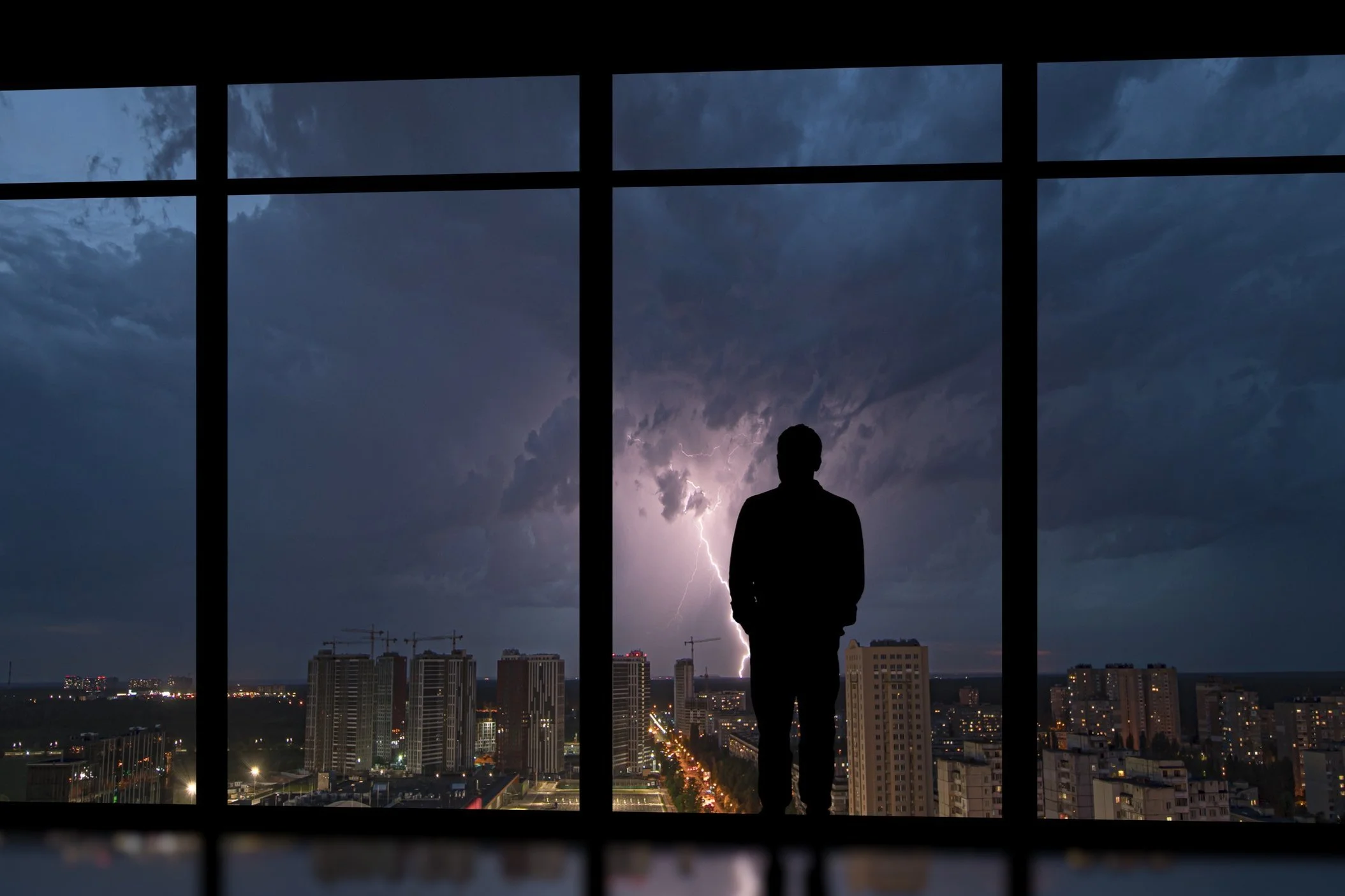 Silhouette of a person standing by a window, observing lightning storm over a city skyline at night.