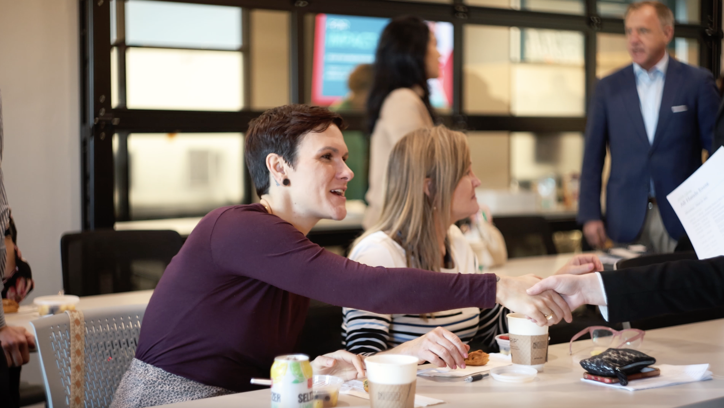 A woman in a maroon top is shaking hands with someone across the table during a meeting or conference. She is smiling and engaged in conversation, with coffee cups and a water bottle on the table. Other attendees are seated nearby, and a man in a blue suit is standing in the background.