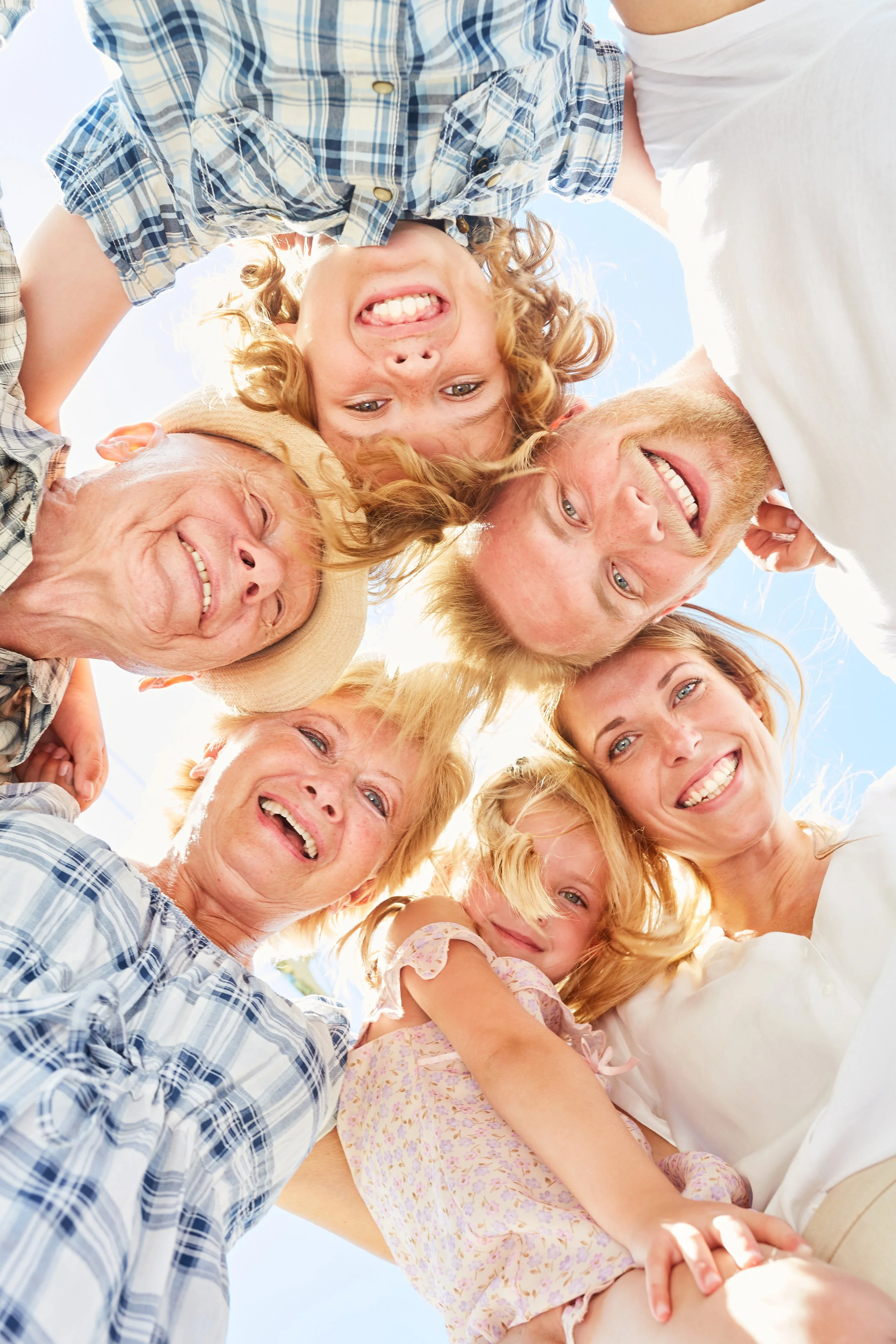 A family of eight, including children and seniors, standing in a circle outdoors, looking down at the camera with smiles under a clear blue sky.