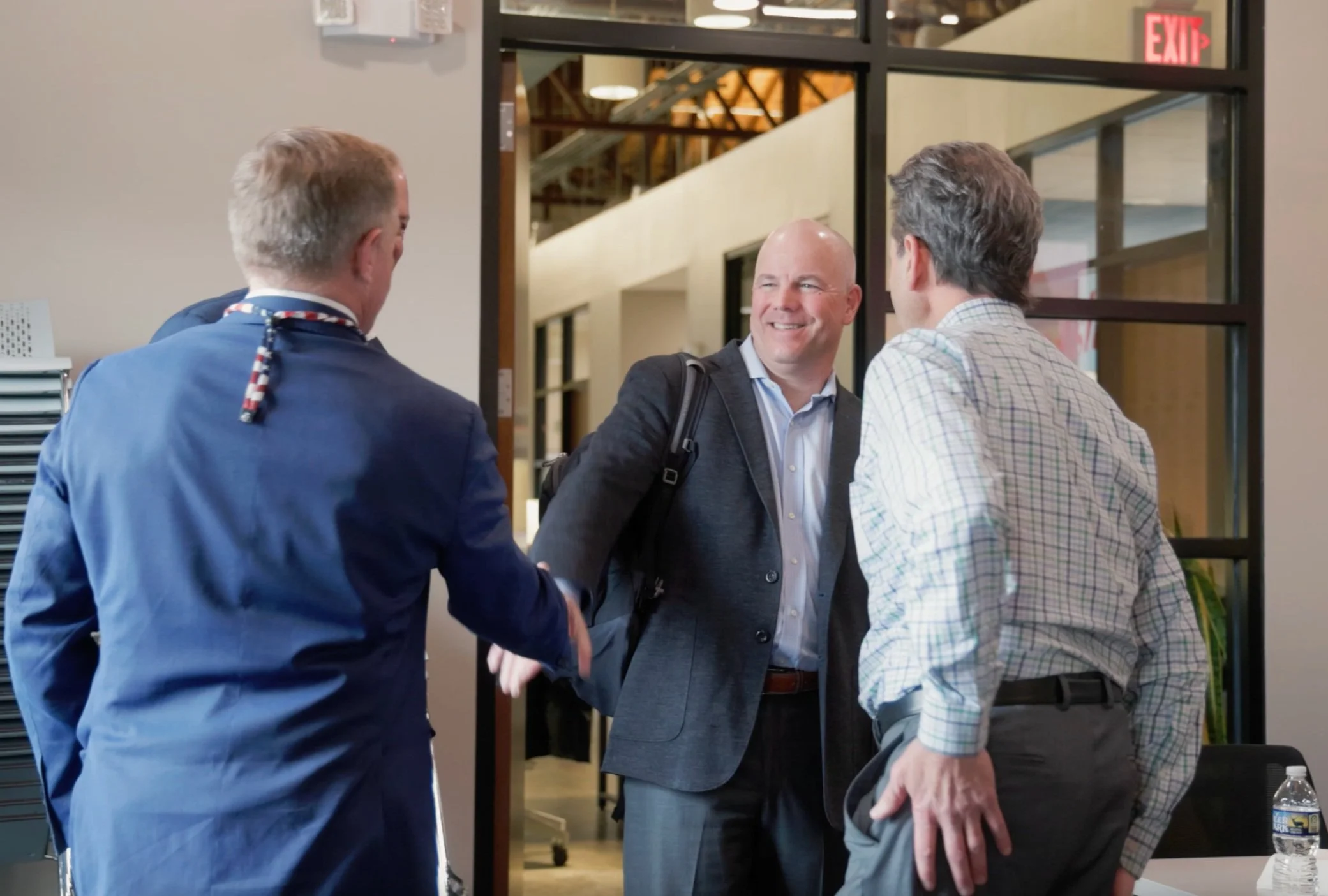Three men in business attire greeting each other and shaking hands inside an office. One man is dressed in a navy blue suit with a colorful scarf, another is dressed in a dark suit with a backpack, and the third is in a checked shirt. They are smiling and engaging in friendly conversation.