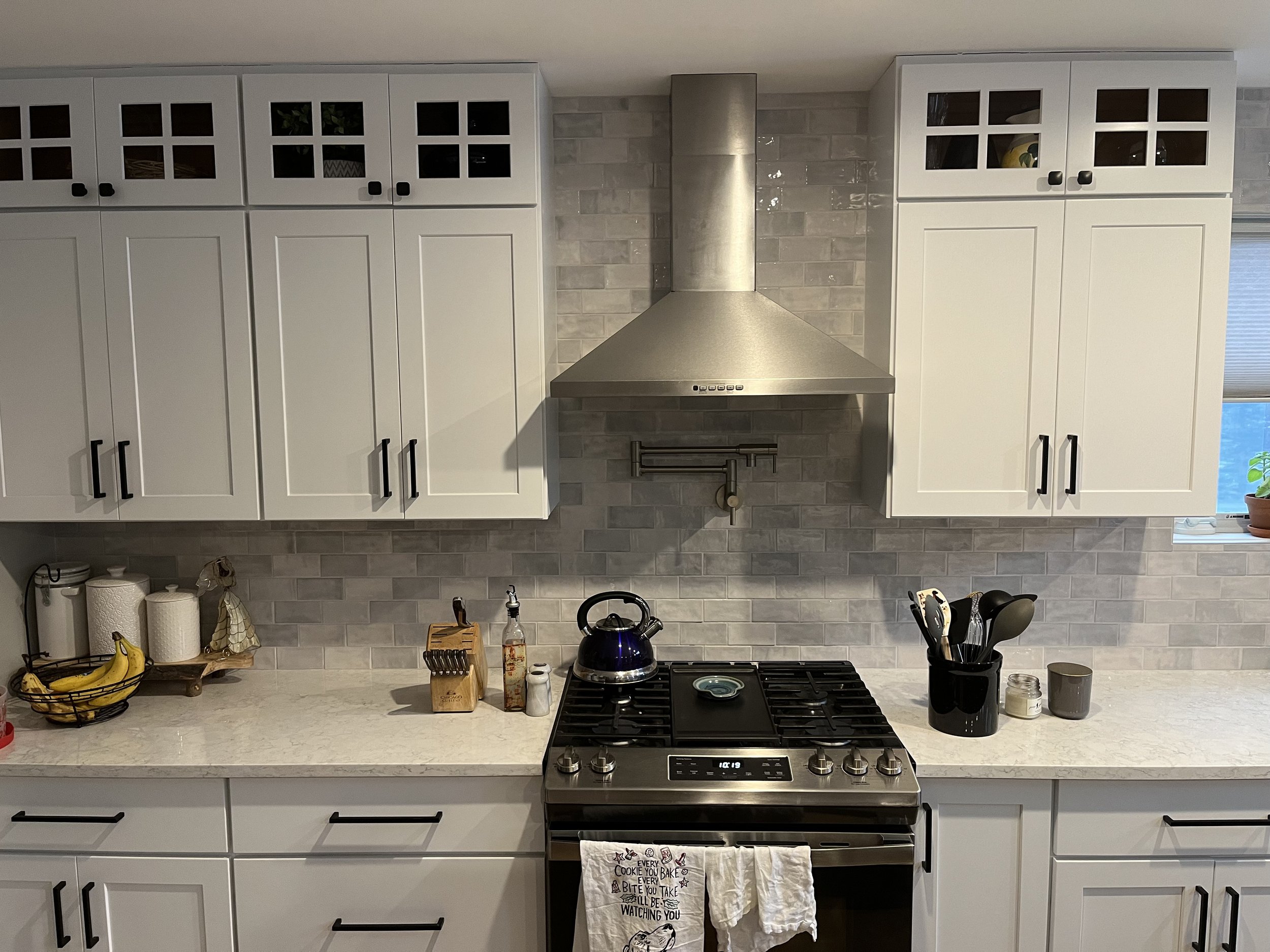 Modern kitchen with white cabinets, stainless steel stove and range hood, gray tile backsplash, black utensils, bananas in a basket, and a small window with a plant on the sill.
