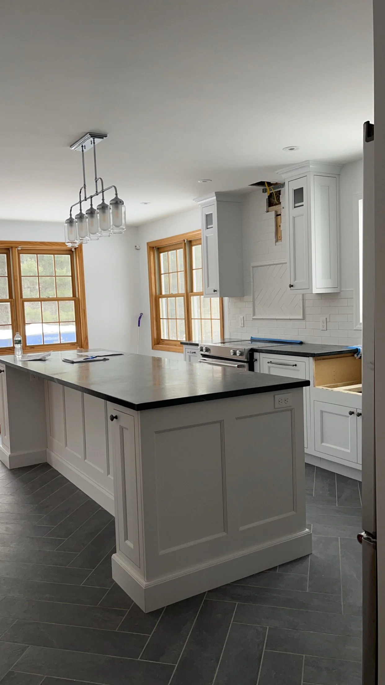 Modern kitchen with white cabinets, a large island with a dark countertop, and windows with wooden frames. Some cabinets are missing doors, and there is an ongoing renovation.
