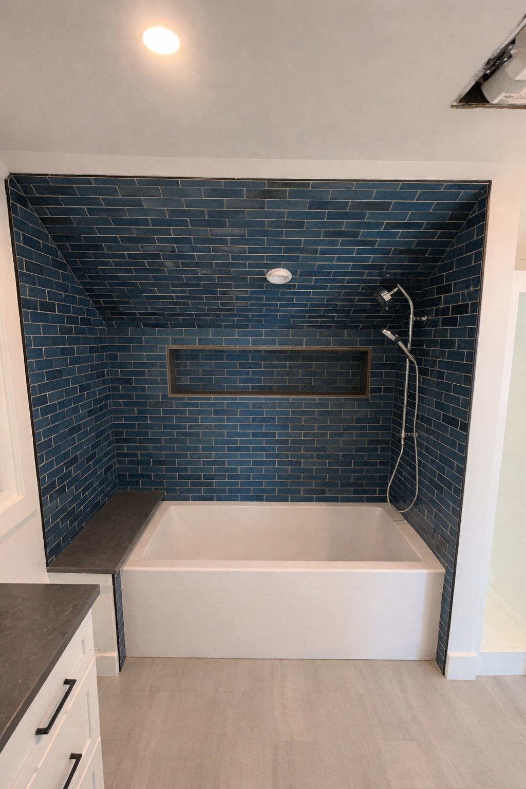 Bathroom with bathtub and blue subway tile in shower area, which has a built-in niche, a showerhead, and a recessed shower seat. Part of a white vanity with black handles is visible on the left, and a ceiling light is turned on.