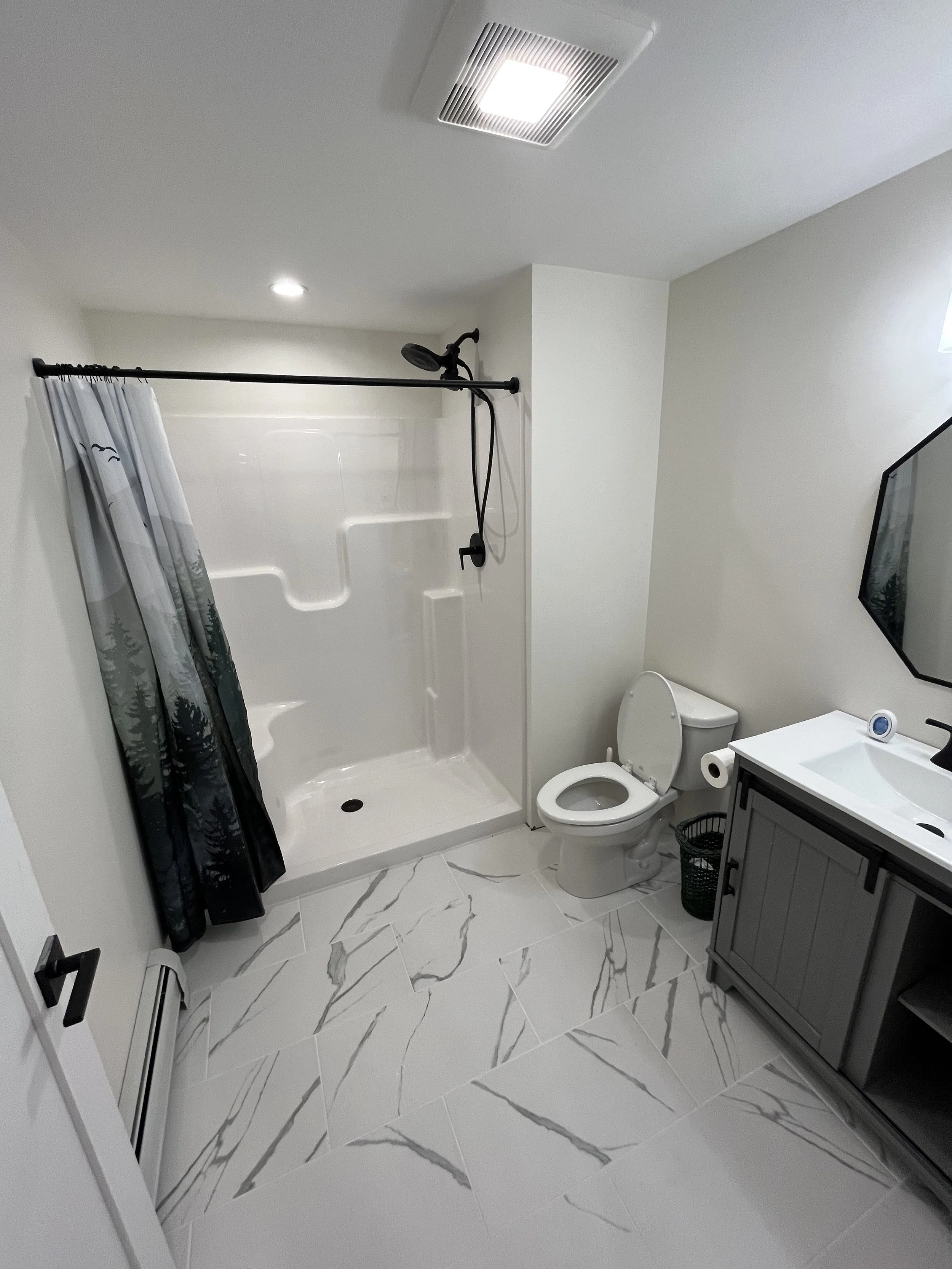 Modern bathroom with white walls and marble-patterned floor tiles, a shower with a black showerhead and curtain, a toilet, and a gray vanity with a sink and black mirror.