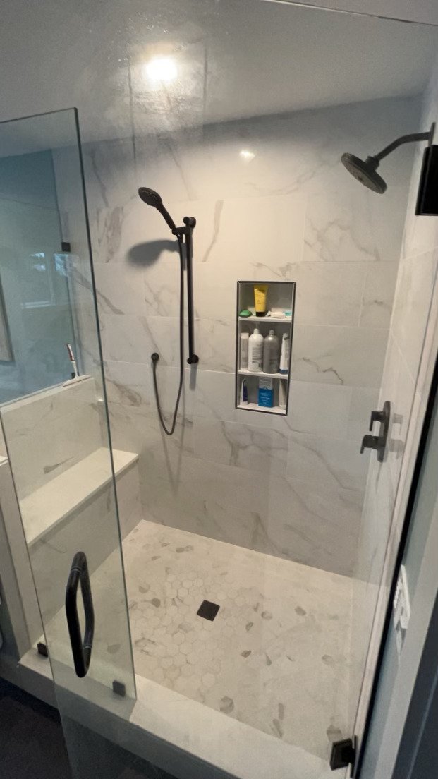 Modern walk-in shower with marble walls and hexagonal tile floor, featuring a built-in shelf with toiletries, black shower fixtures, and a glass door.