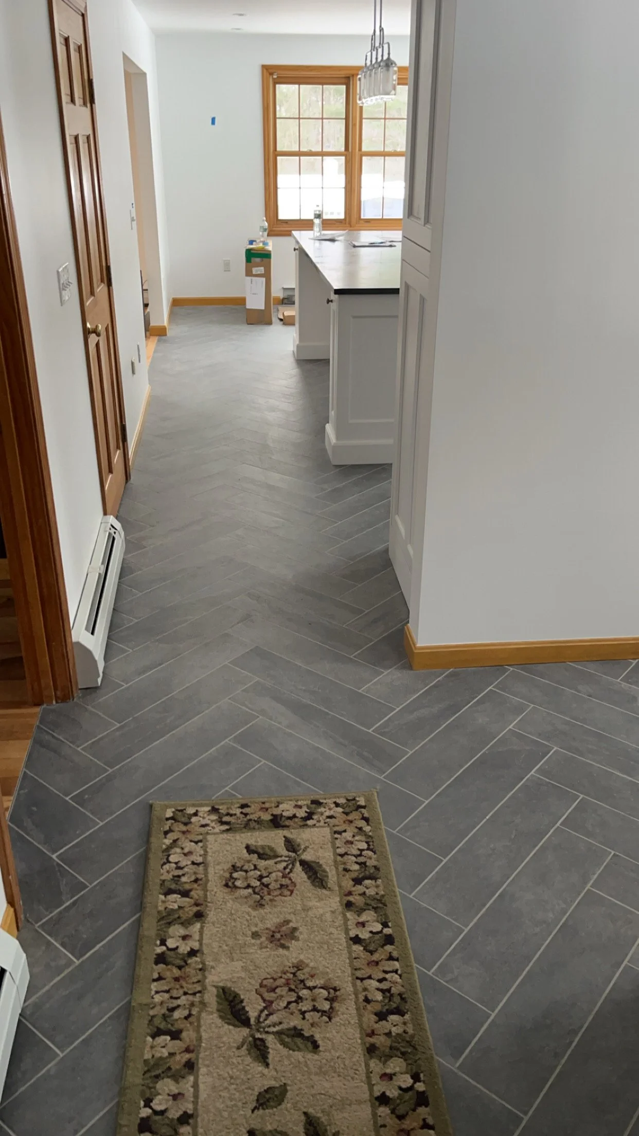 View of a kitchen with gray herringbone tile flooring, white cabinets, and a large window with wooden trim. There is a small floral rug and some boxes near the window, suggesting ongoing setup or renovation.