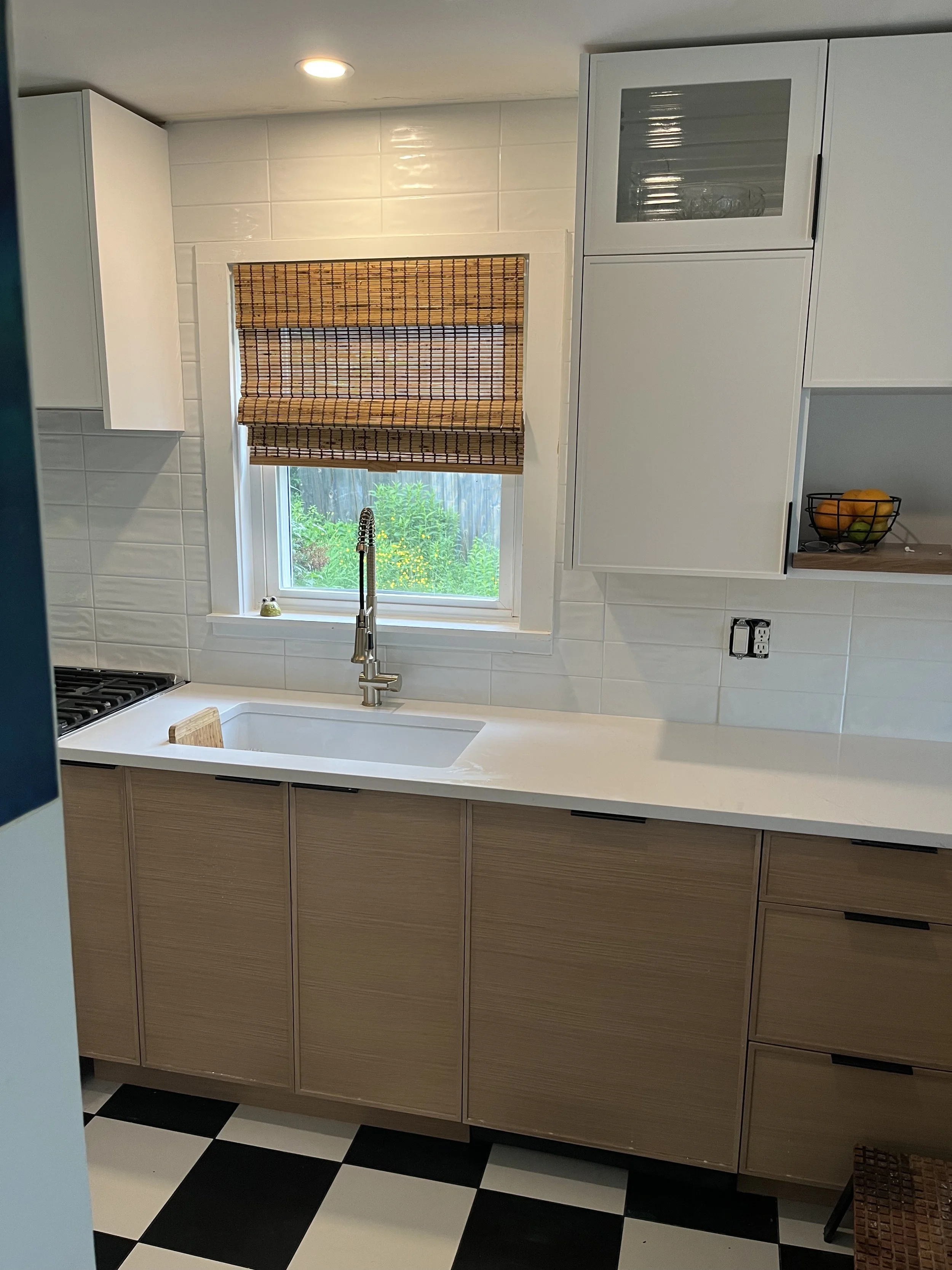 Kitchen with white cabinets, a window with bamboo blinds, checkered black and white floor, and a window view of greenery.