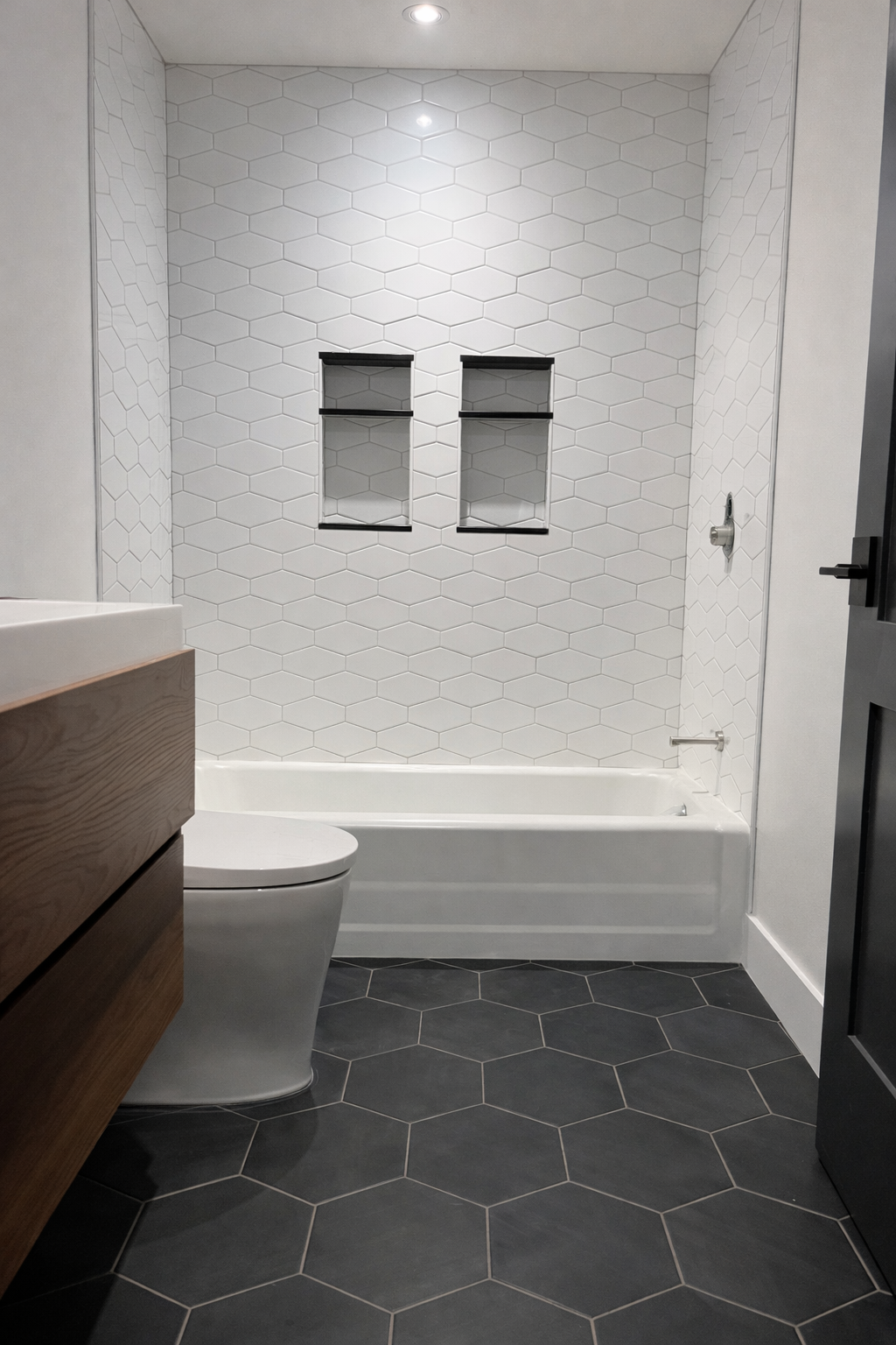 Modern bathroom with white hexagonal tile on the walls, dark hexagonal tile on the floor, a white bathtub, a wall with two recessed shelves, and a partial view of a toilet and a wooden vanity.