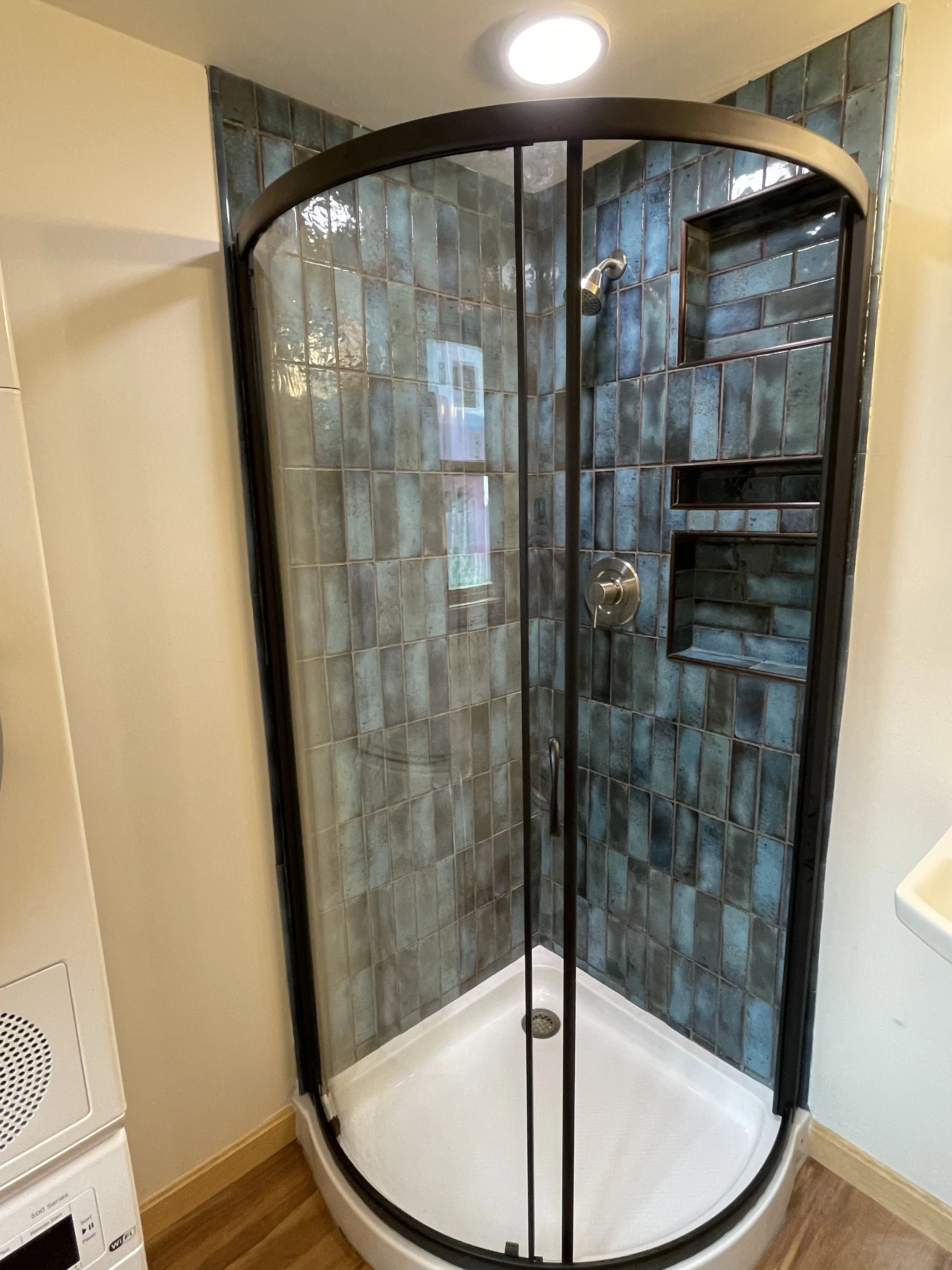 Curved glass shower stall with dark metal frame, blue-green tile interior, built-in shelving, and a showerhead.