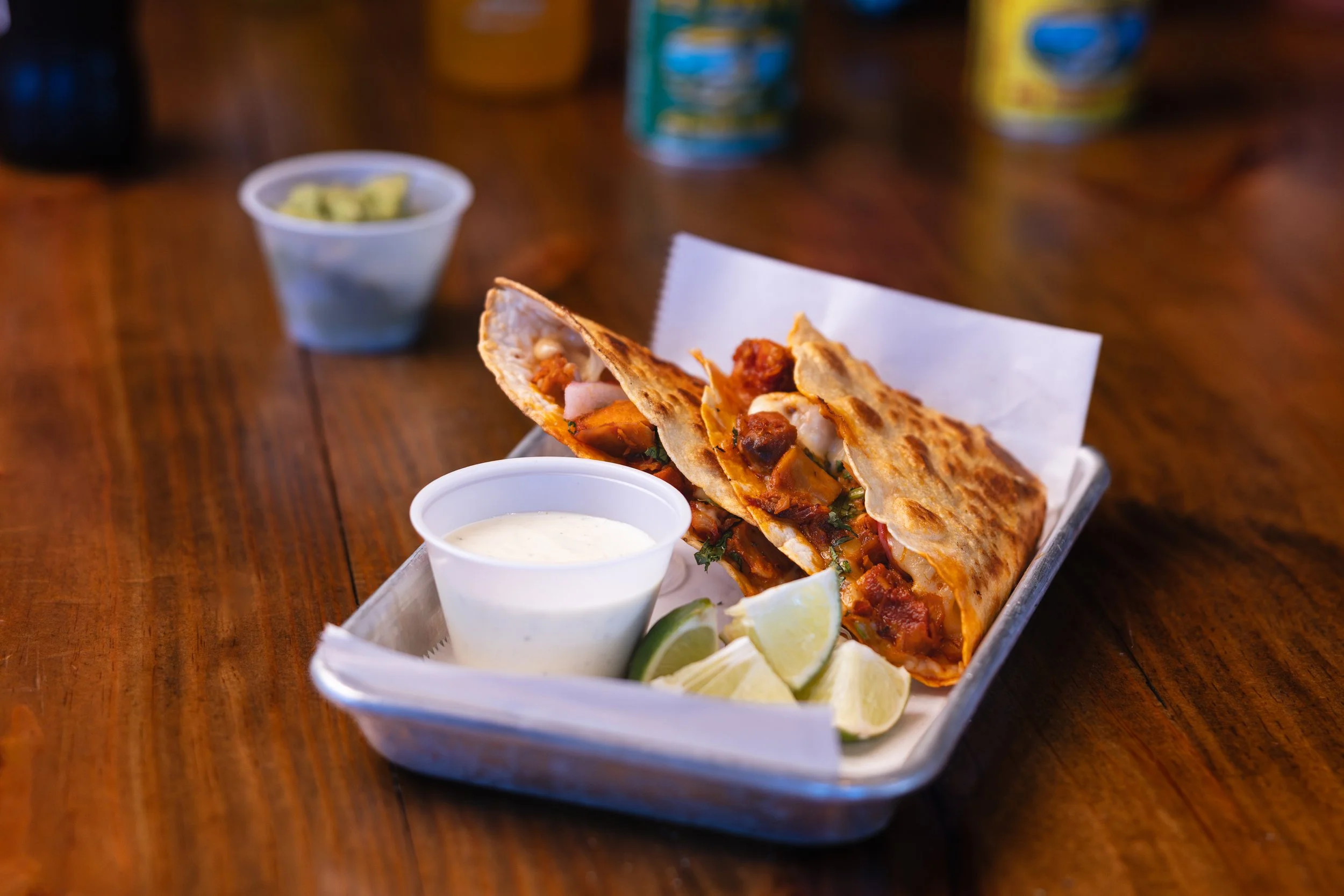 A plate of two chili-filled burritos with lime wedges and two cups of creamy white sauce, with a small cup of guacamole in the background on a wooden table.